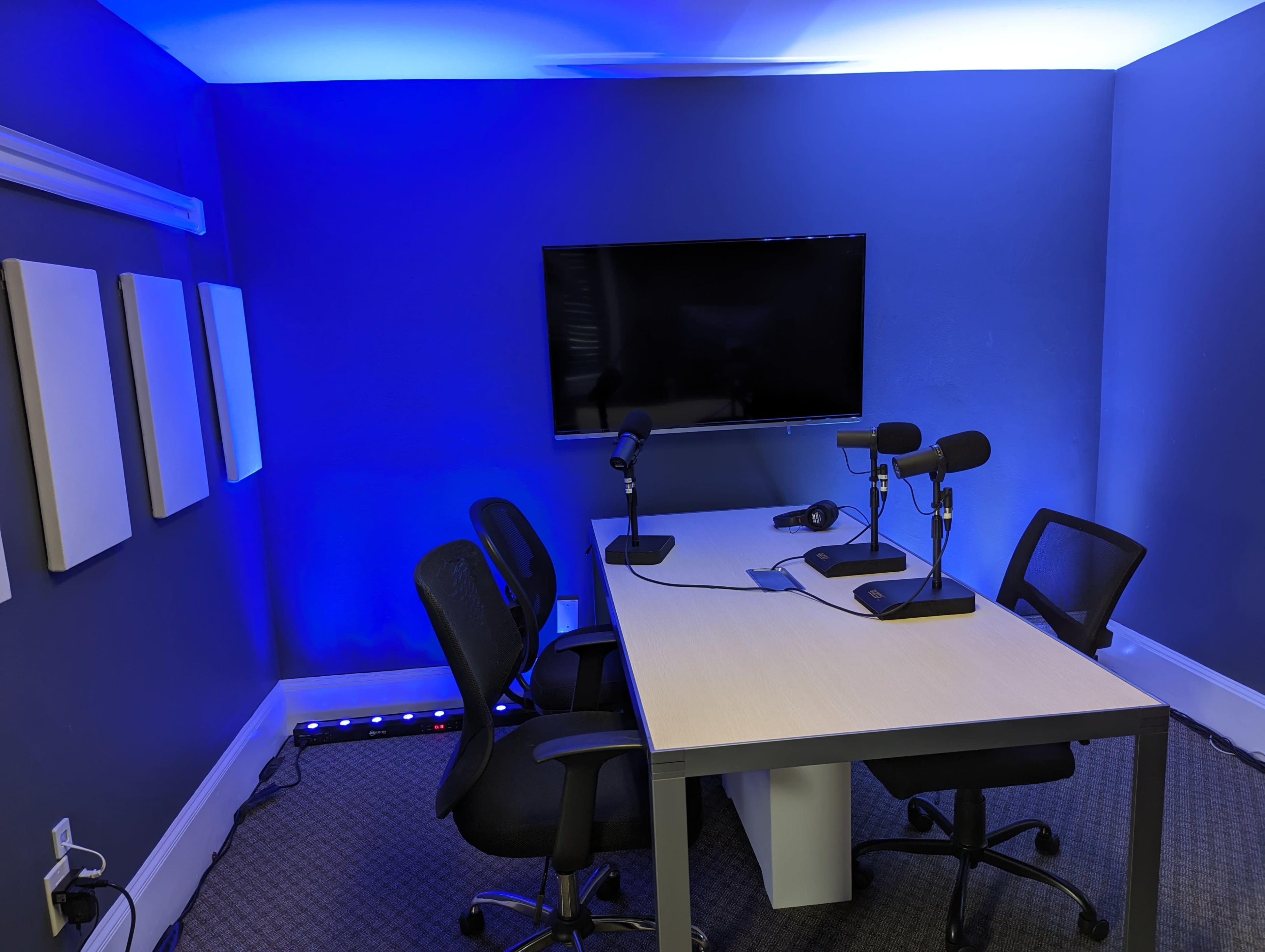 A recording studio features a table with two microphones and four chairs under blue lighting and a large screen on the wall.