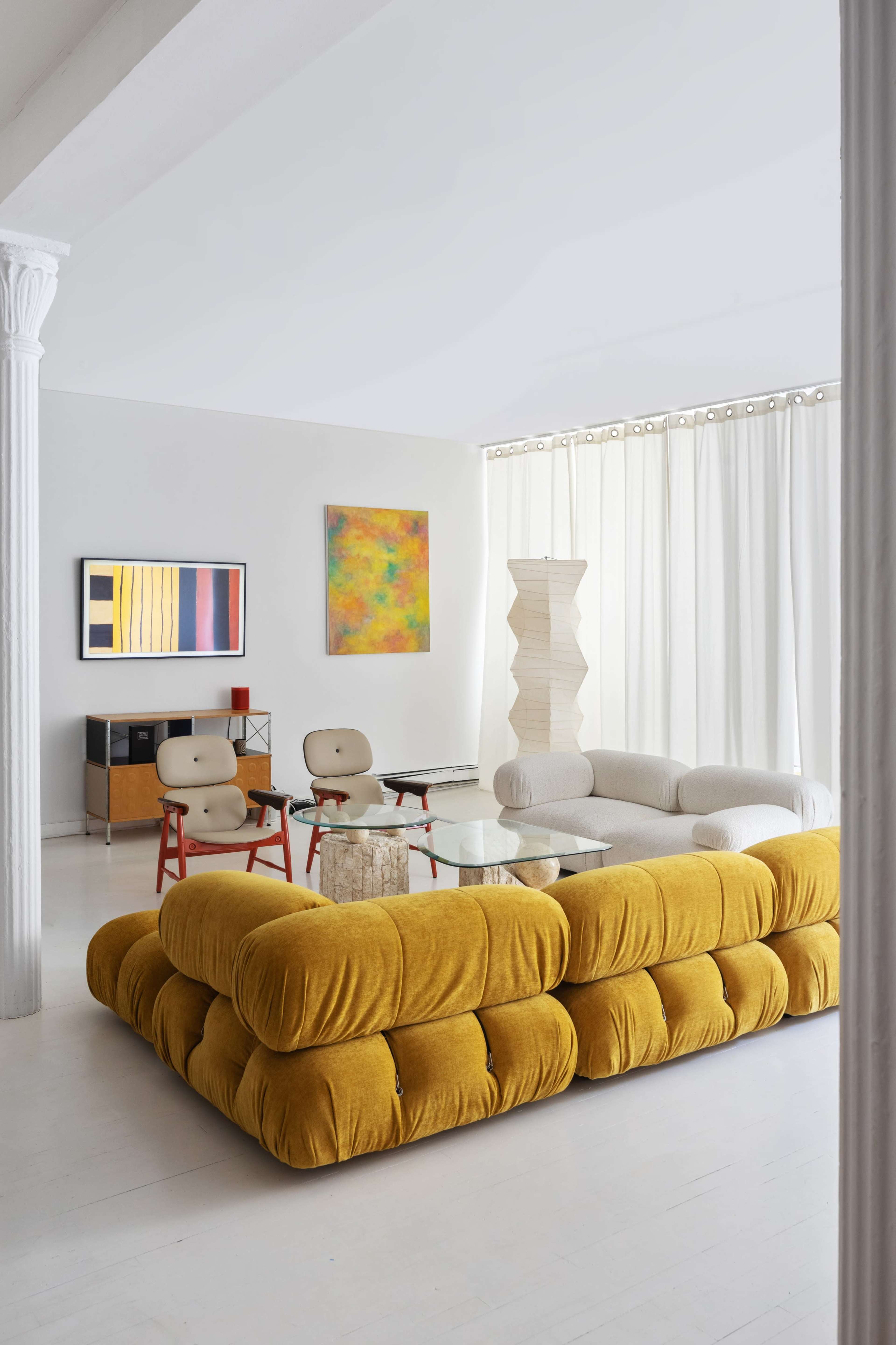 The image shows a modern living room featuring a large mustard-colored sectional sofa, a glass coffee table, and minimalist decor with colorful artwork on the walls.