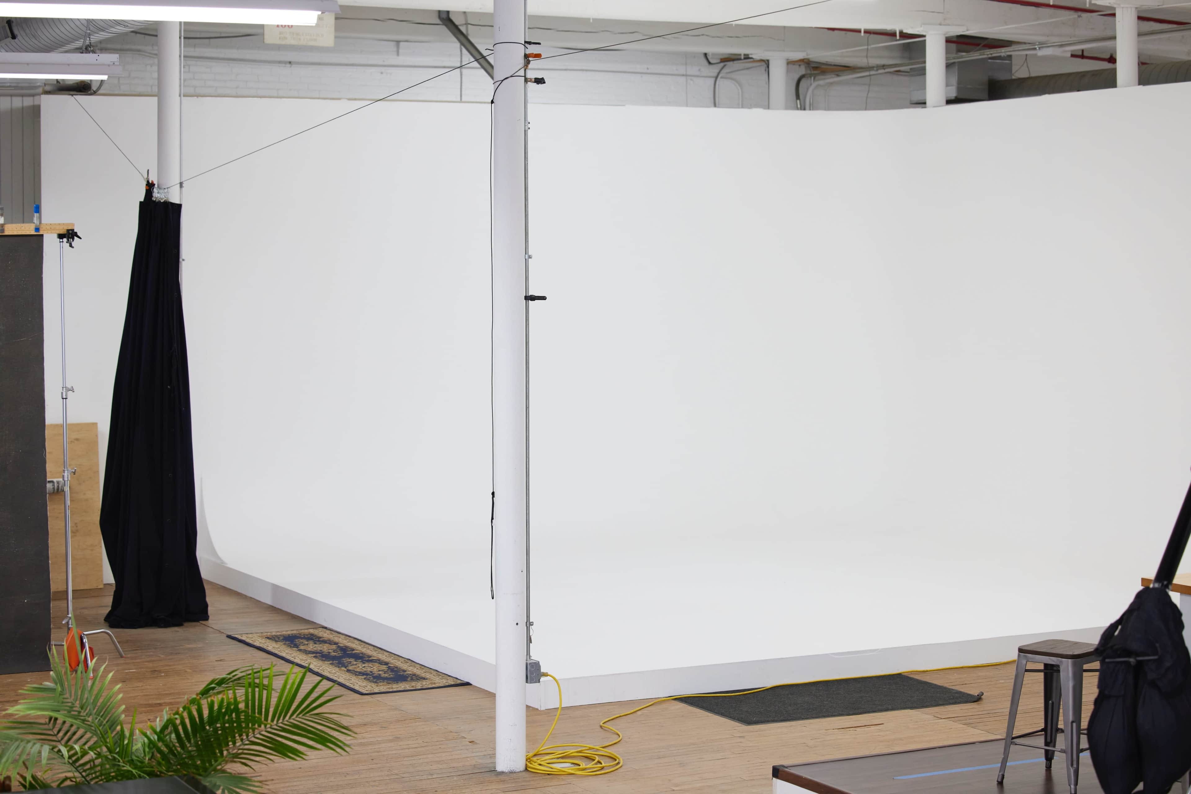 A photography studio with a white backdrop, a wooden floor, and a few scattered props, including a plant and a cable on the ground.