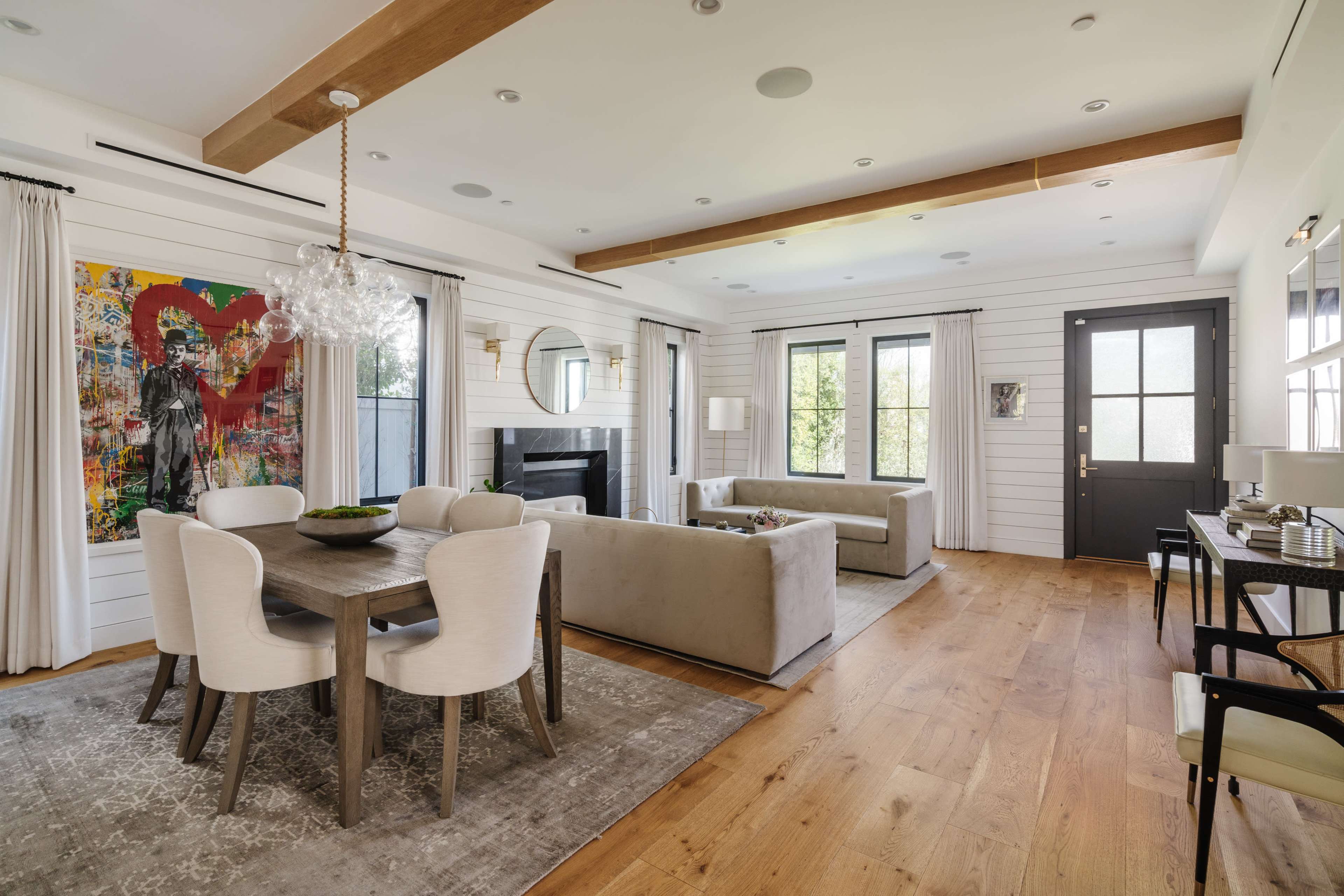 A modern living area with a beige sectional sofa, a dining table with white chairs, a colorful wall art piece, and large windows.