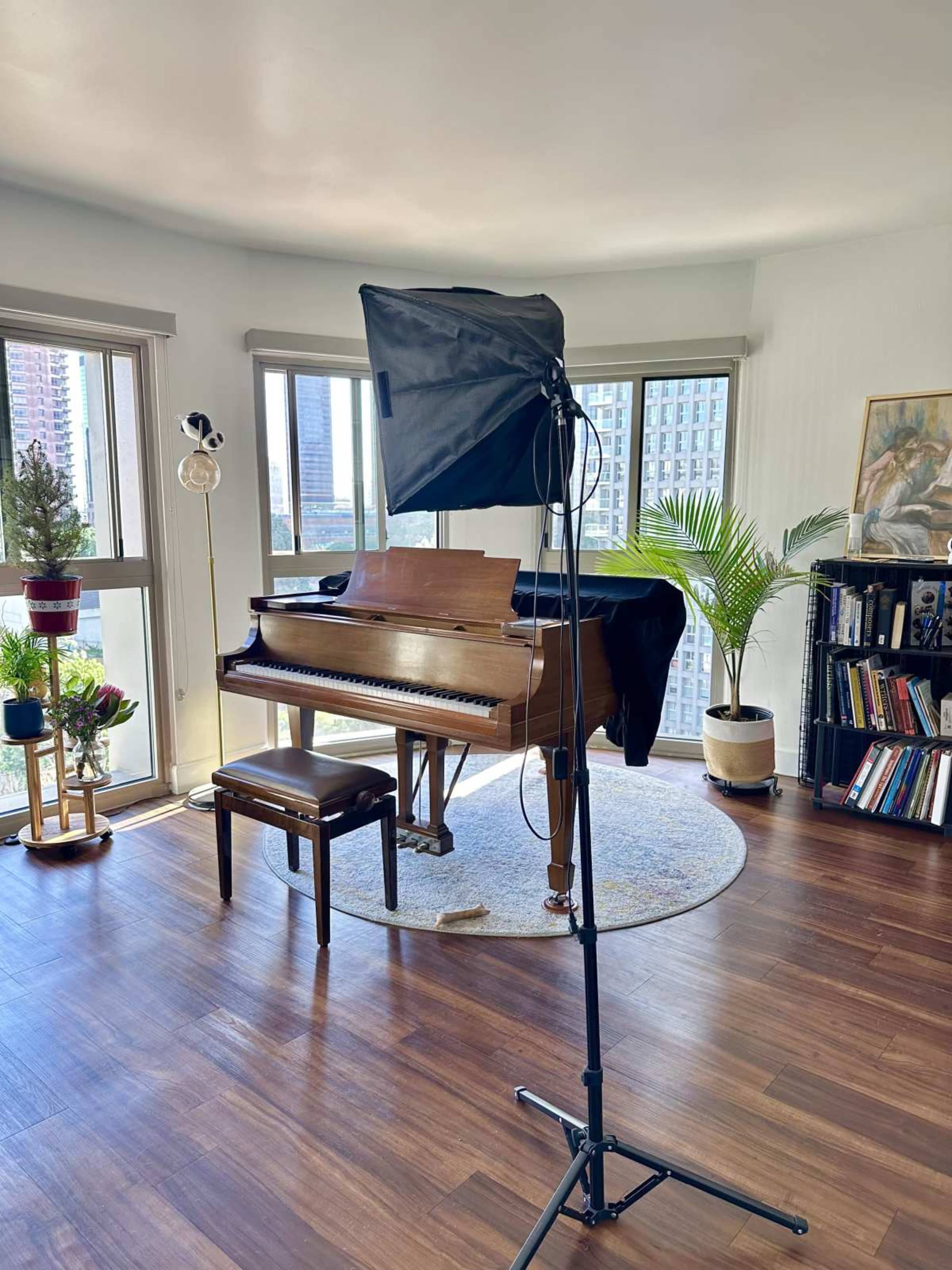 The image shows a well-lit room featuring a grand piano with a bench, a black backdrop on a stand, and large windows offering a view of a city skyline.