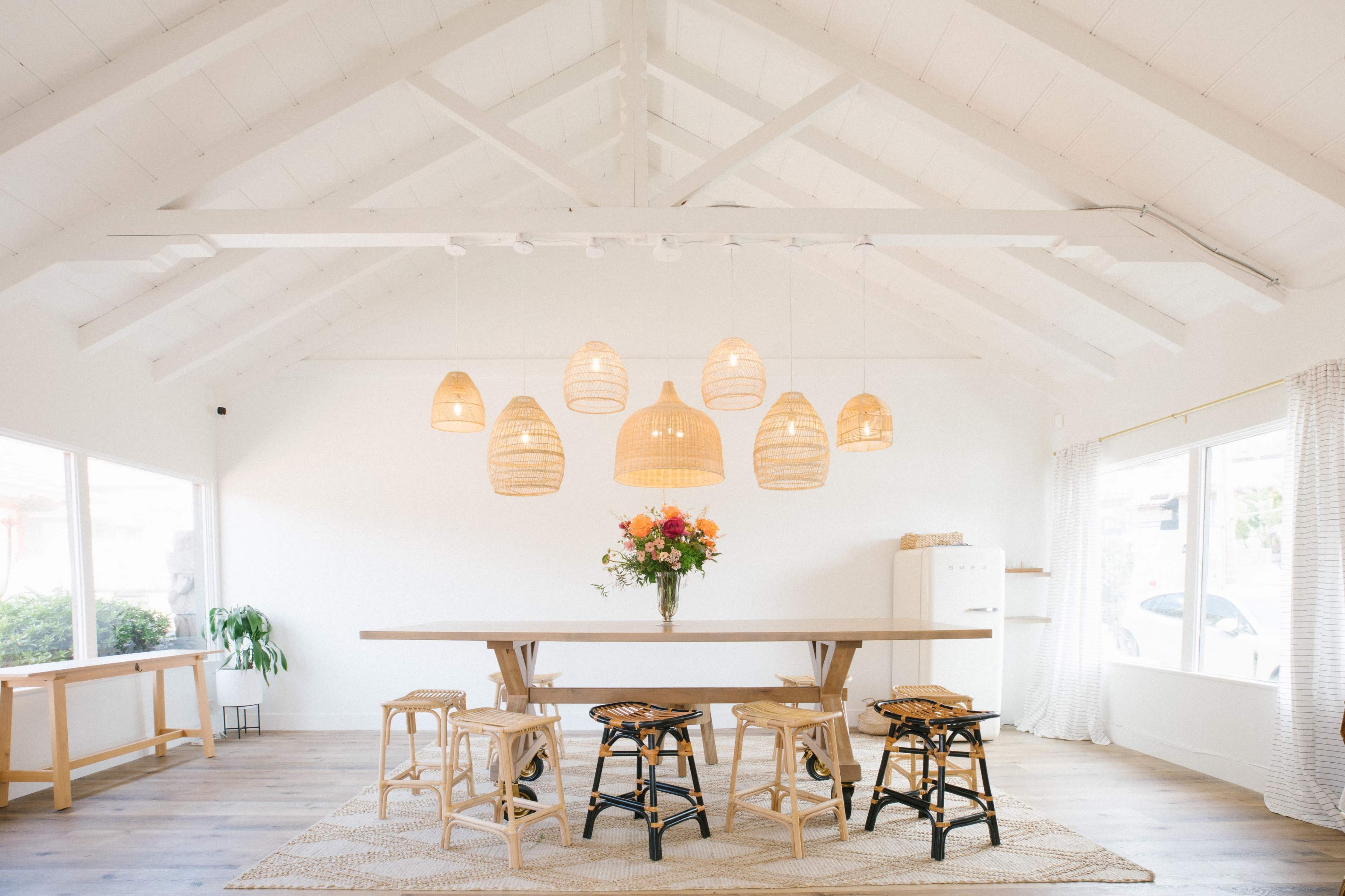 A bright, airy interior features a large wooden dining table surrounded by stools, with overhead rattan light fixtures and a floral arrangement centerpiece.