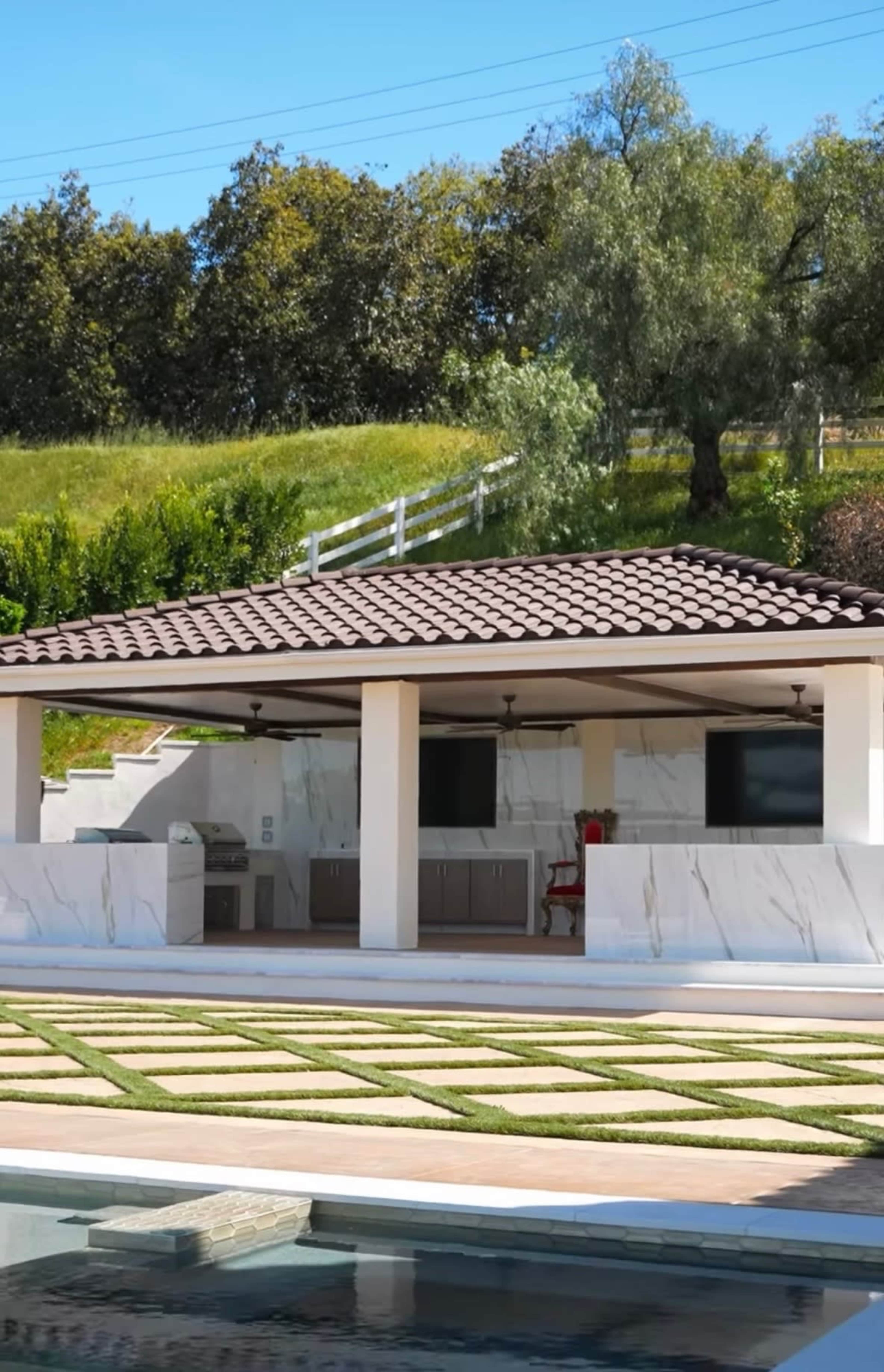The image shows an outdoor kitchen area with a covered pavilion, surrounded by a landscaped yard and a swimming pool.