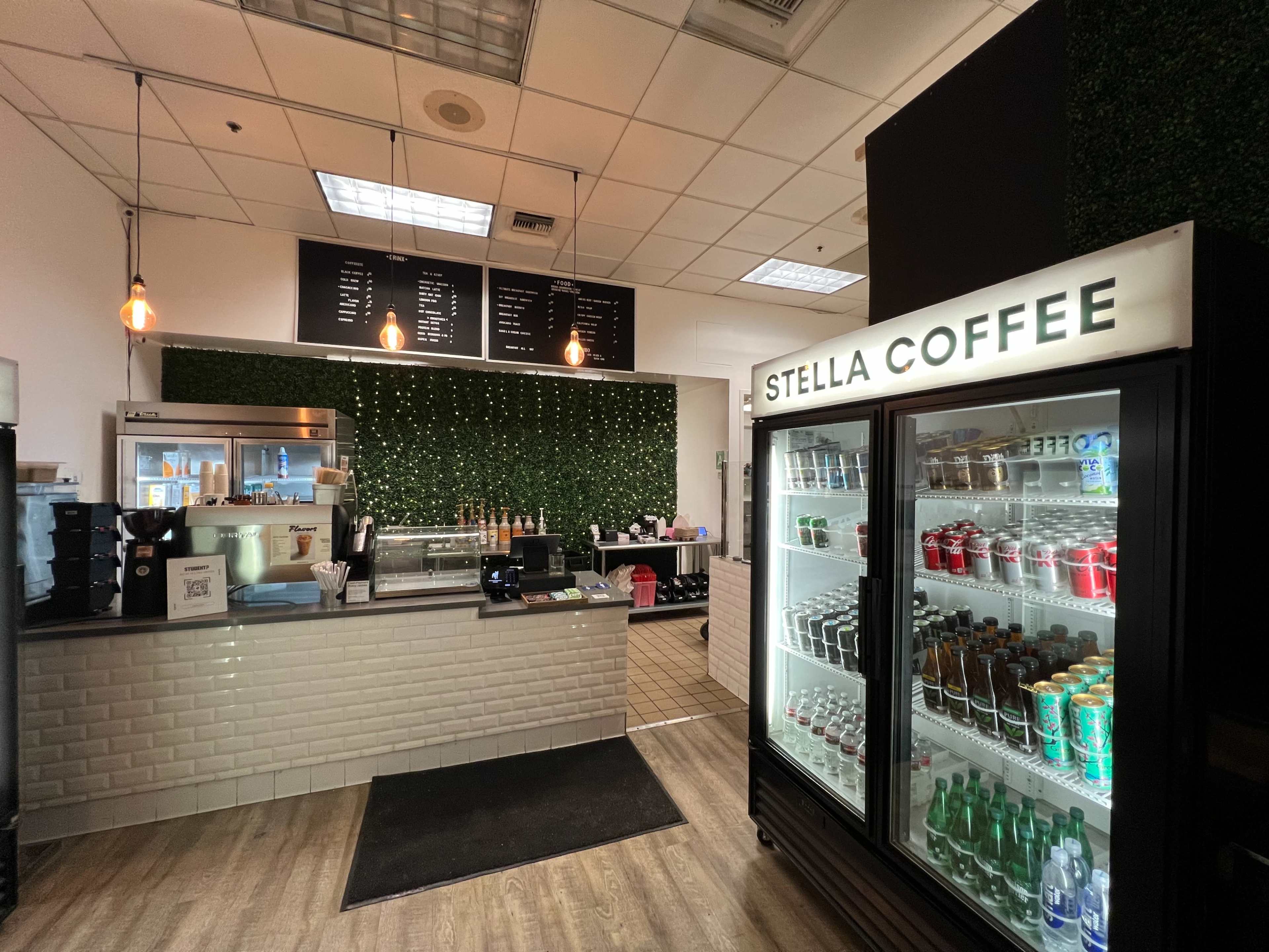 The image shows a modern coffee shop interior with a counter, beverage refrigerator, and a green wall adorned with string lights.
