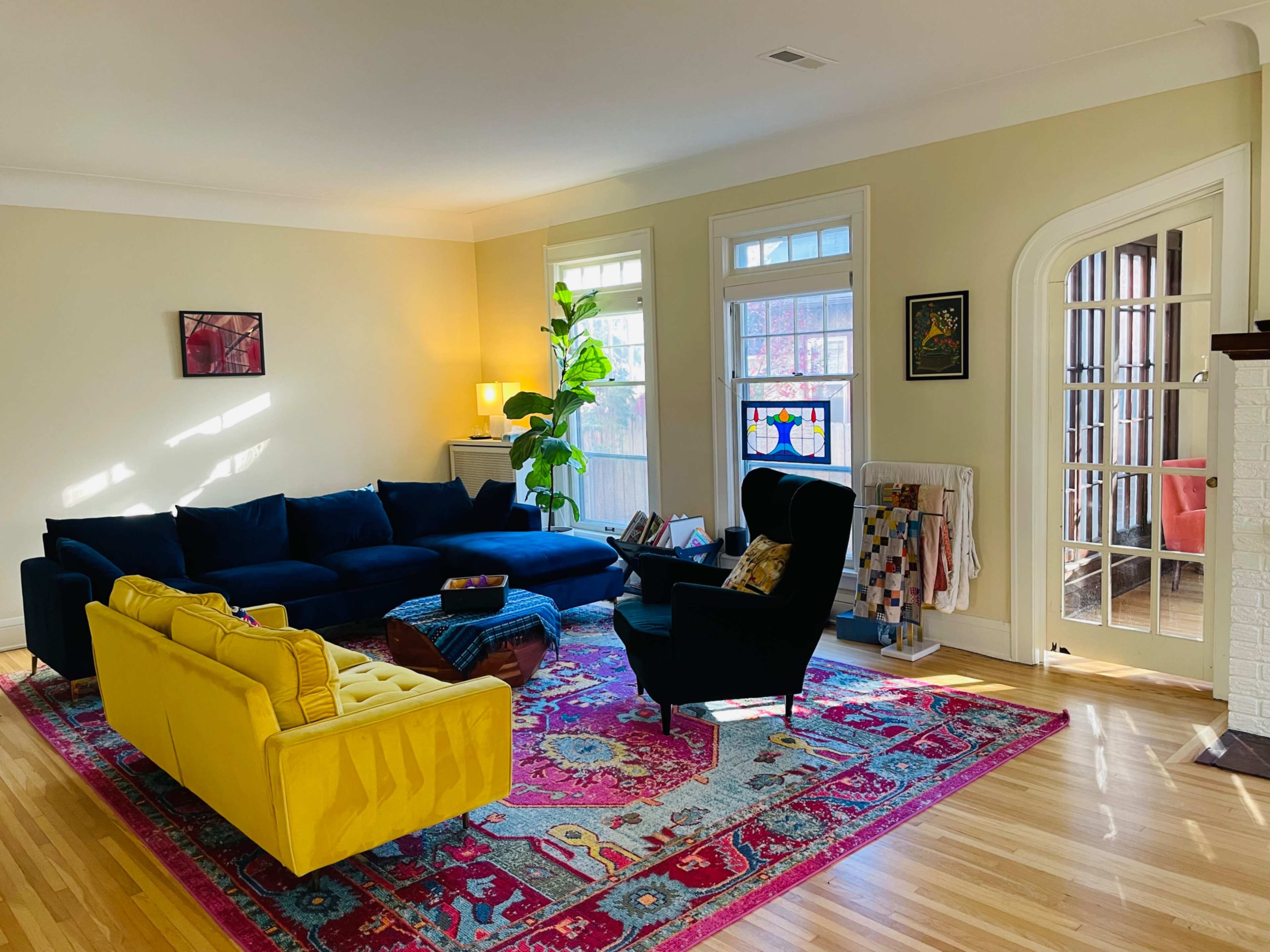 A bright living room features a blue sectional sofa, a yellow couch, and a colorful area rug, with large windows allowing natural light to fill the space.