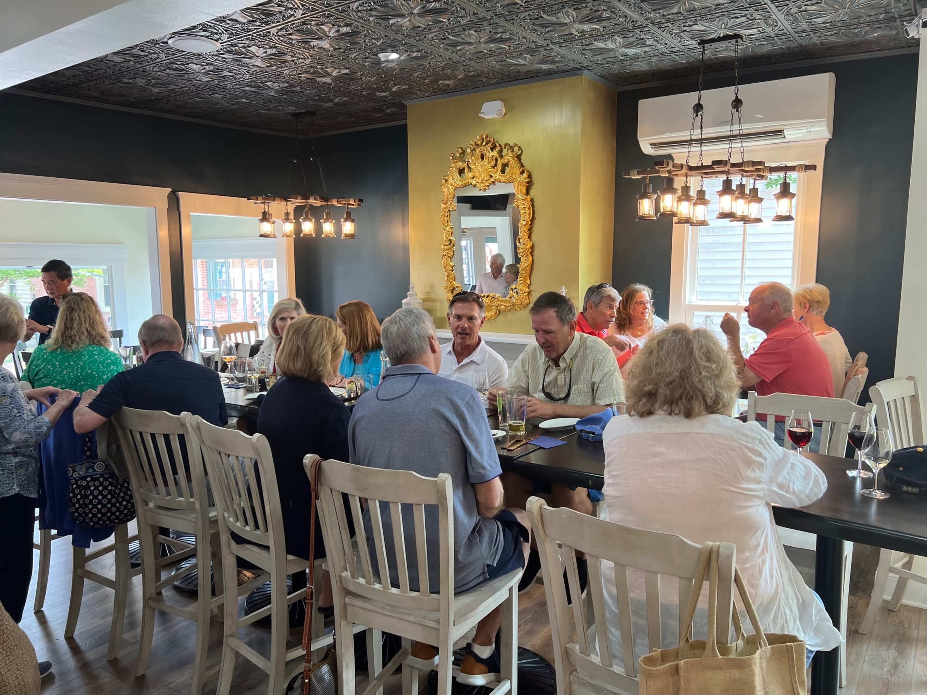 A group of people sits around a large table in a restaurant, engaging in conversation while others are seen in the background.