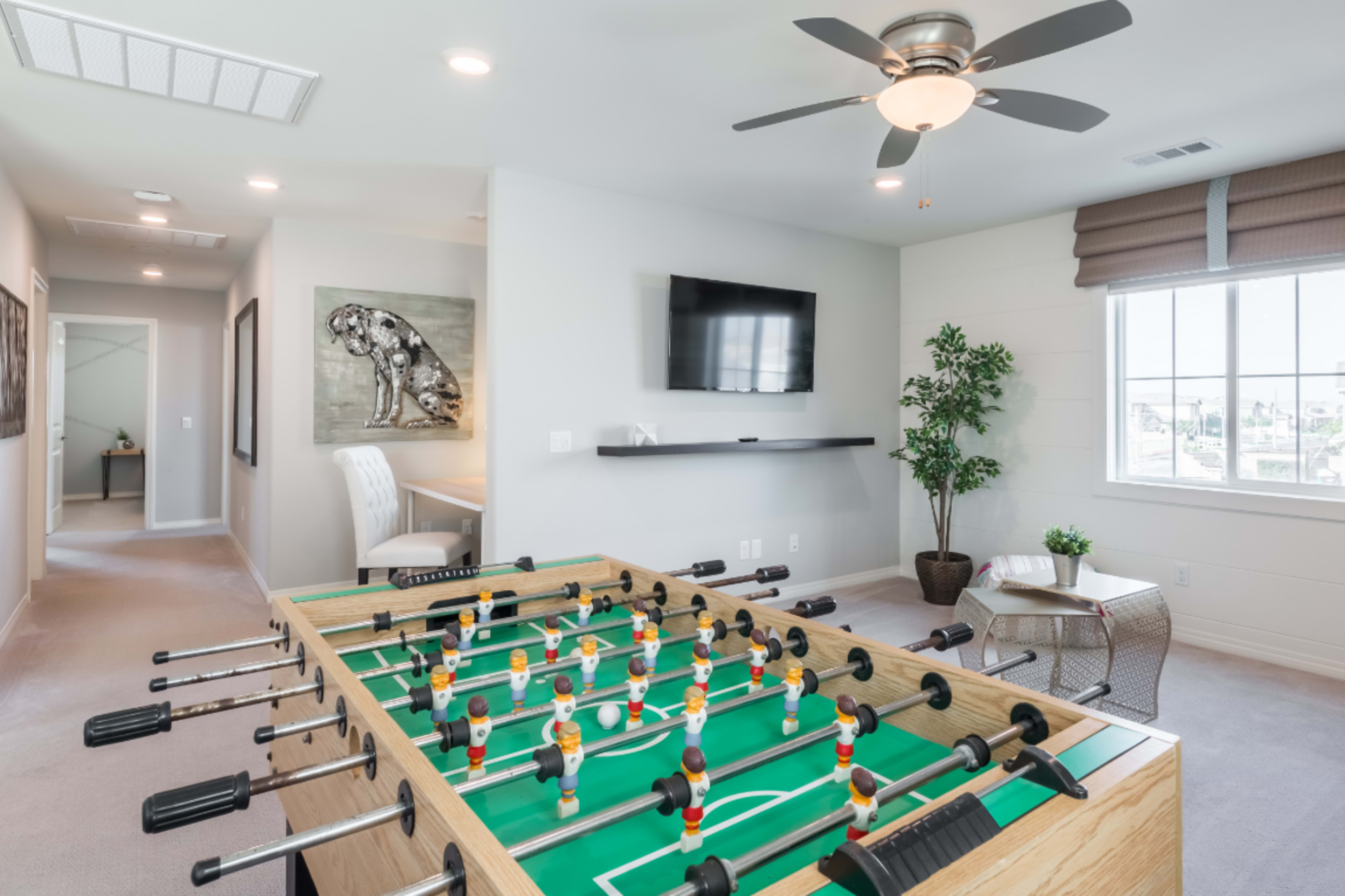 A foosball table occupies the center of a well-lit room featuring a television mounted on the wall and a window with natural light.