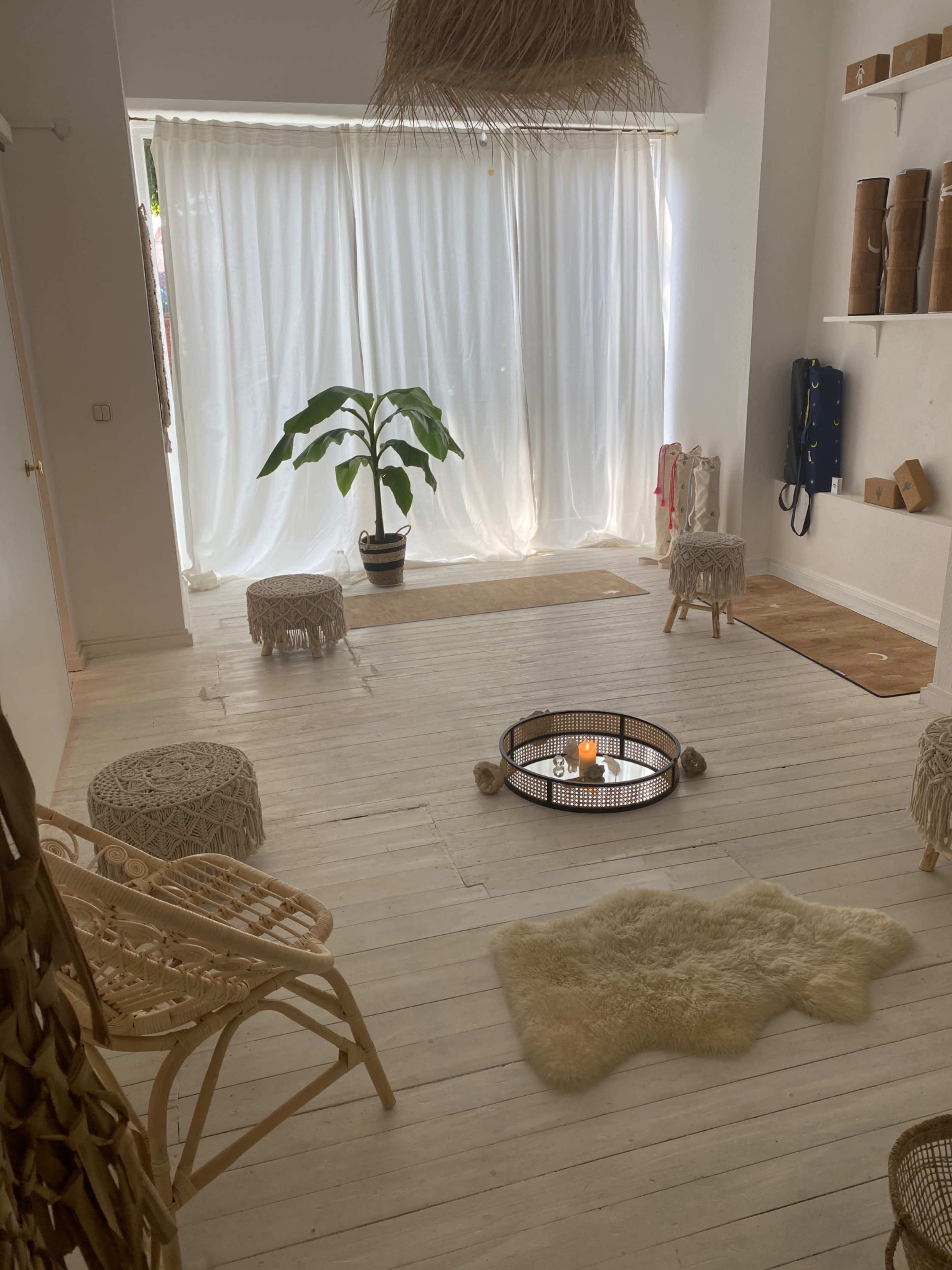 A bright, minimalist room features a large plant, a circular fire pit, woven furniture, and light-colored wooden floors with sheer curtains letting in natural light.