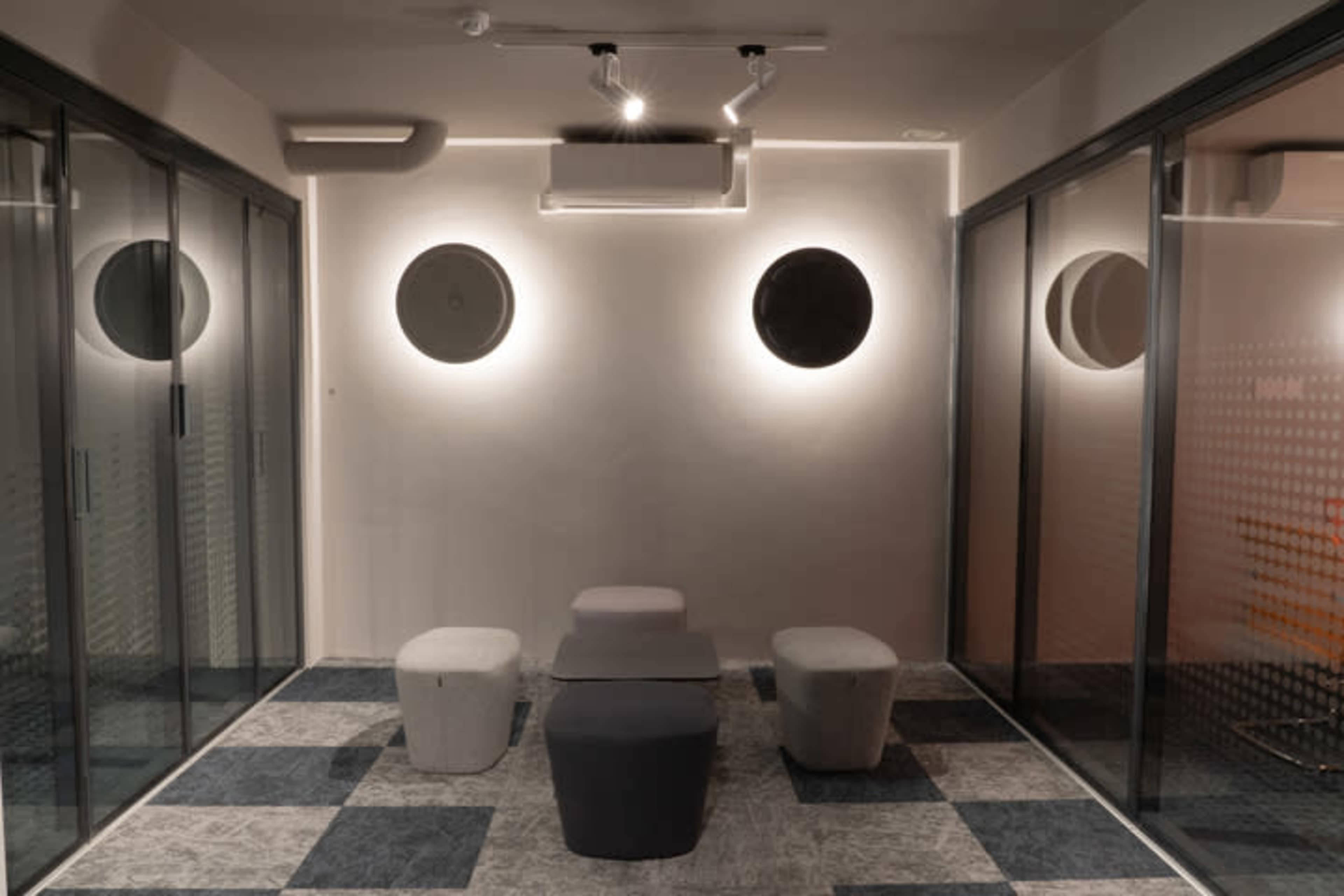 A modern waiting area with three low tables and circular wall lights, enclosed by glass partitions.