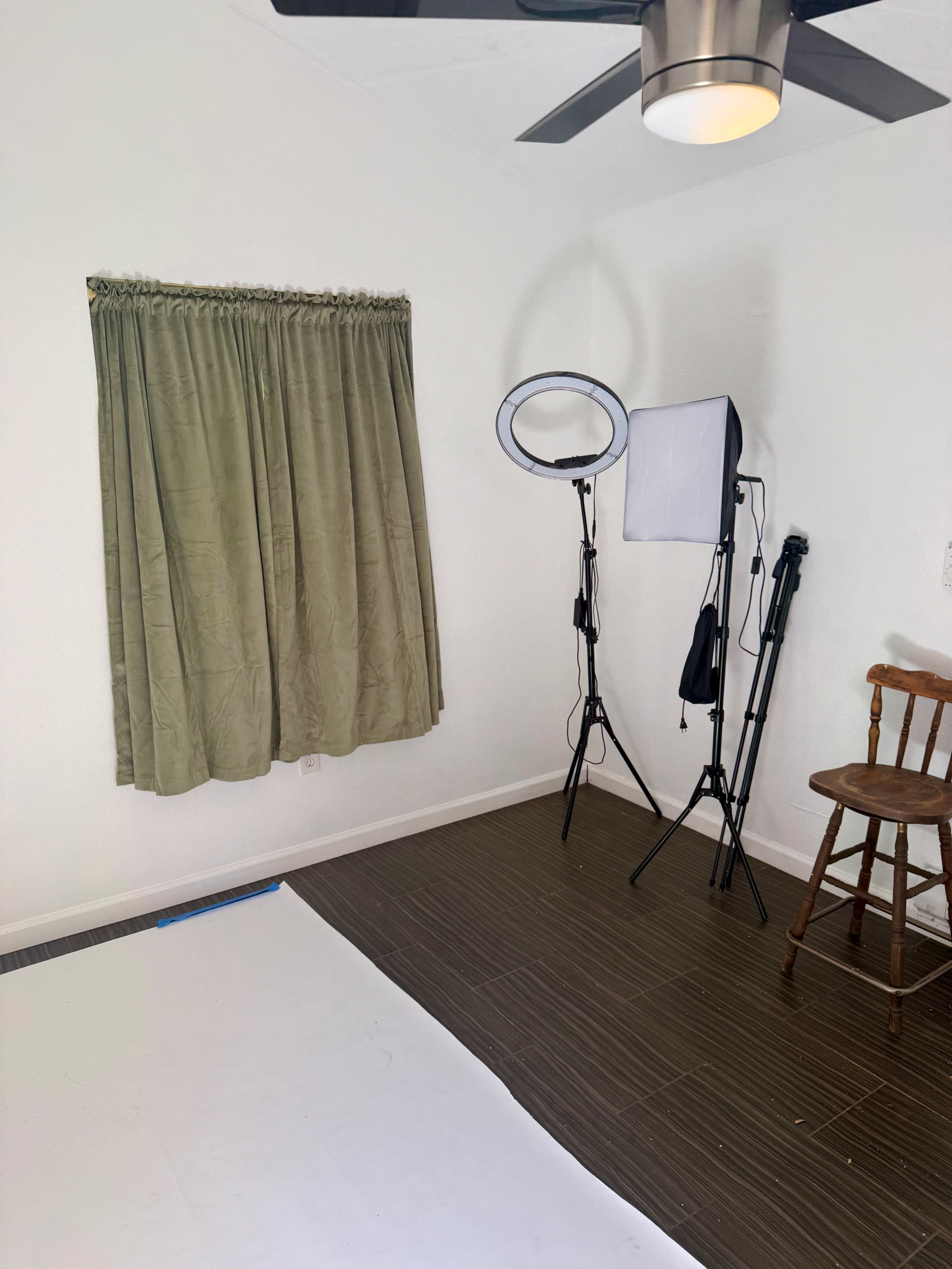 The image shows a small, well-lit room with a curtain-covered window, a ring light on a tripod, a softbox light on a stand, and a wooden chair on a brown floor.
