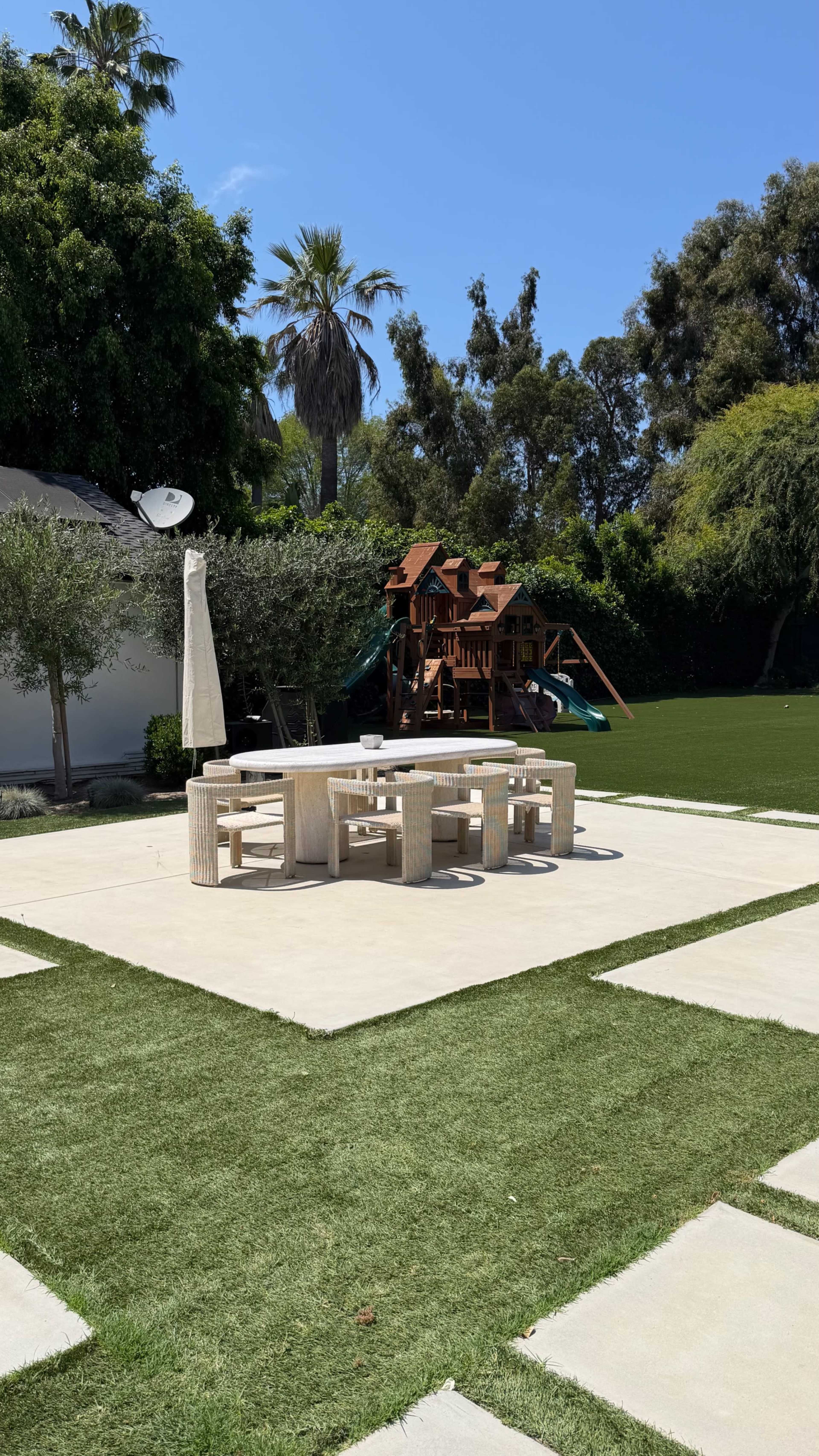 A patio features a circular table with multiple chairs in a landscaped backyard that includes a play structure and palm trees.