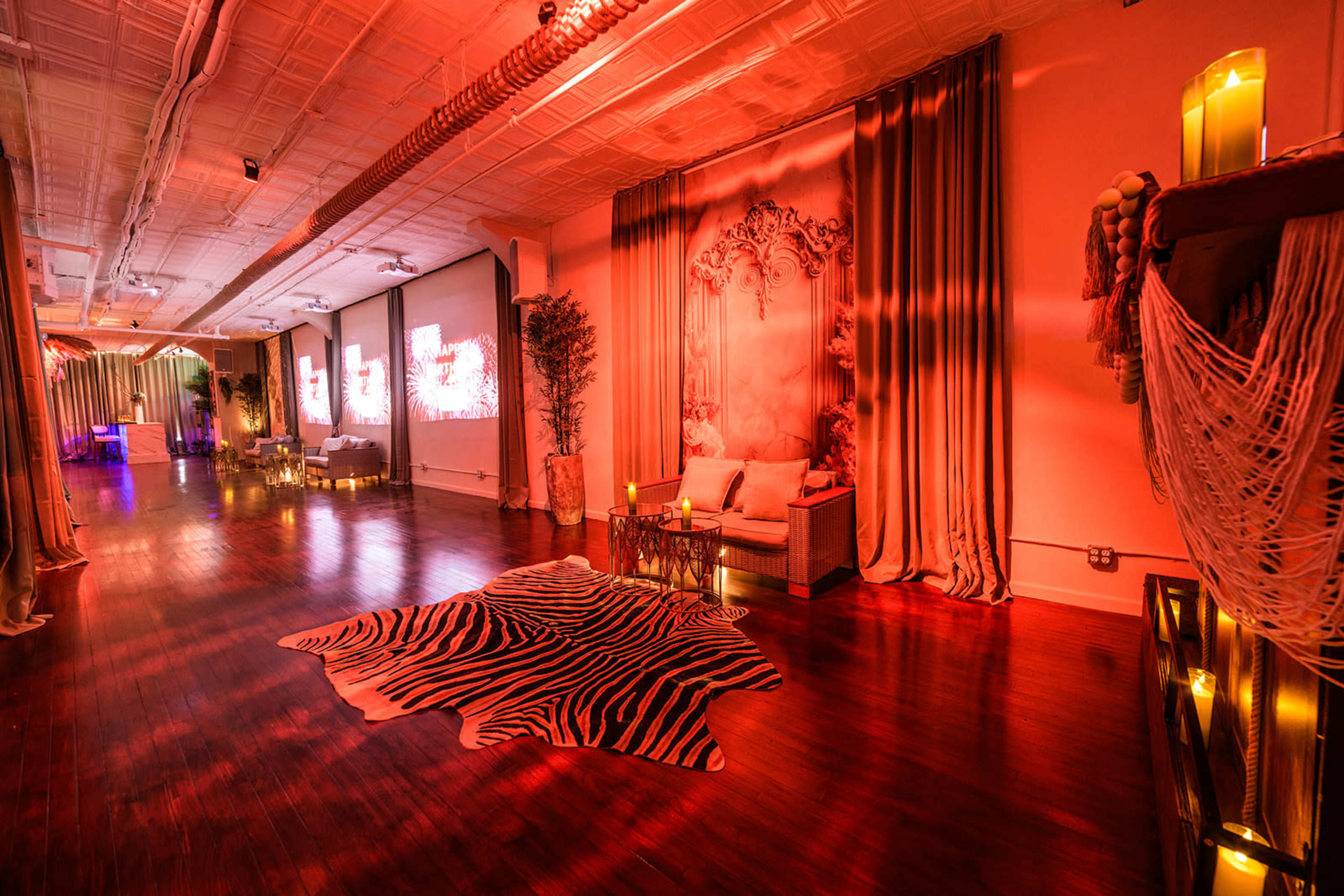 The image shows a warmly lit event space with a zebra-striped rug, decorative curtains, and soft seating arrangements alongside flickering candles.