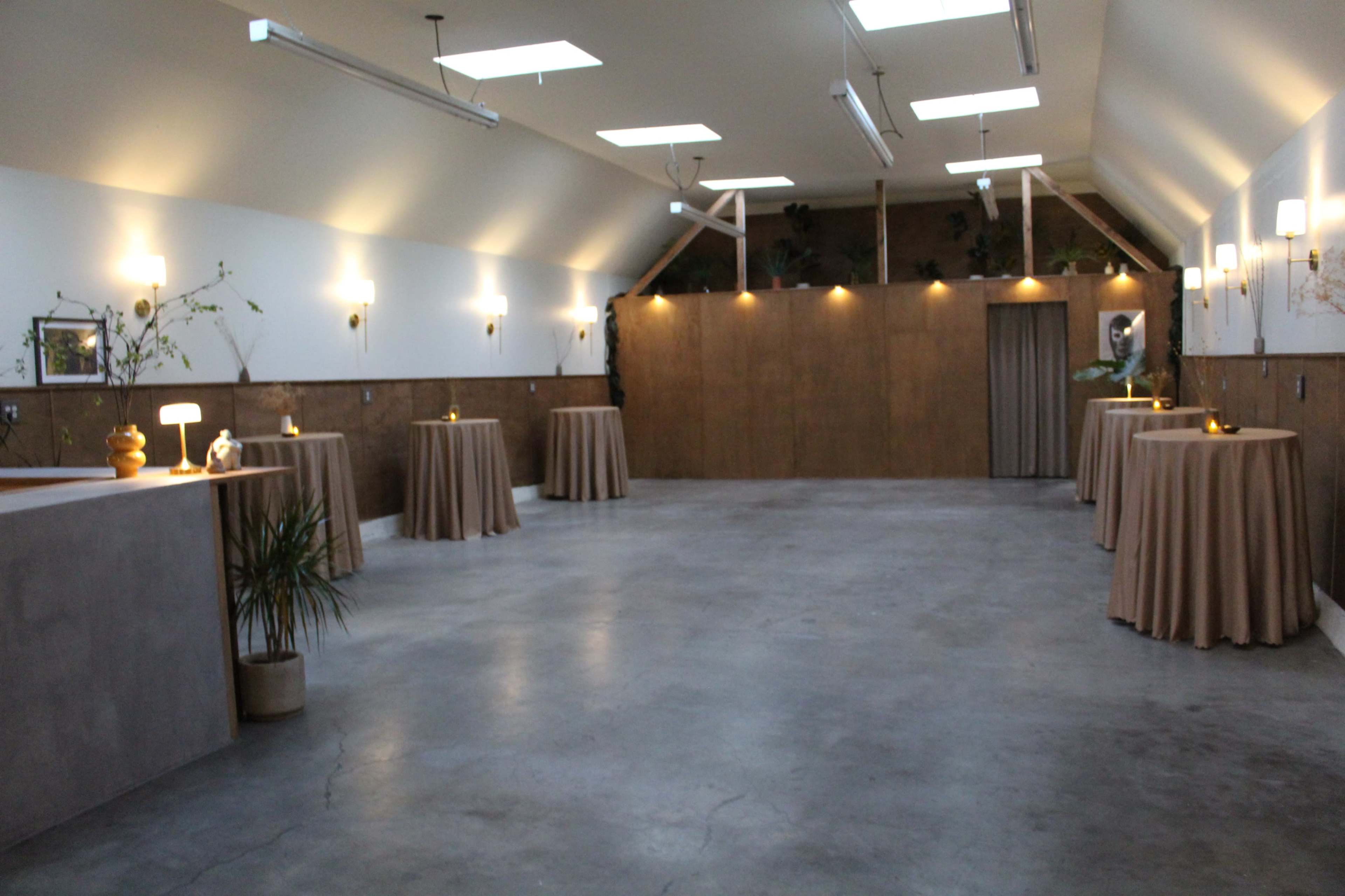 The image shows an empty event space with concrete floors, tall ceilings, and several round tables covered with beige tablecloths, illuminated by soft wall and ceiling lighting.
