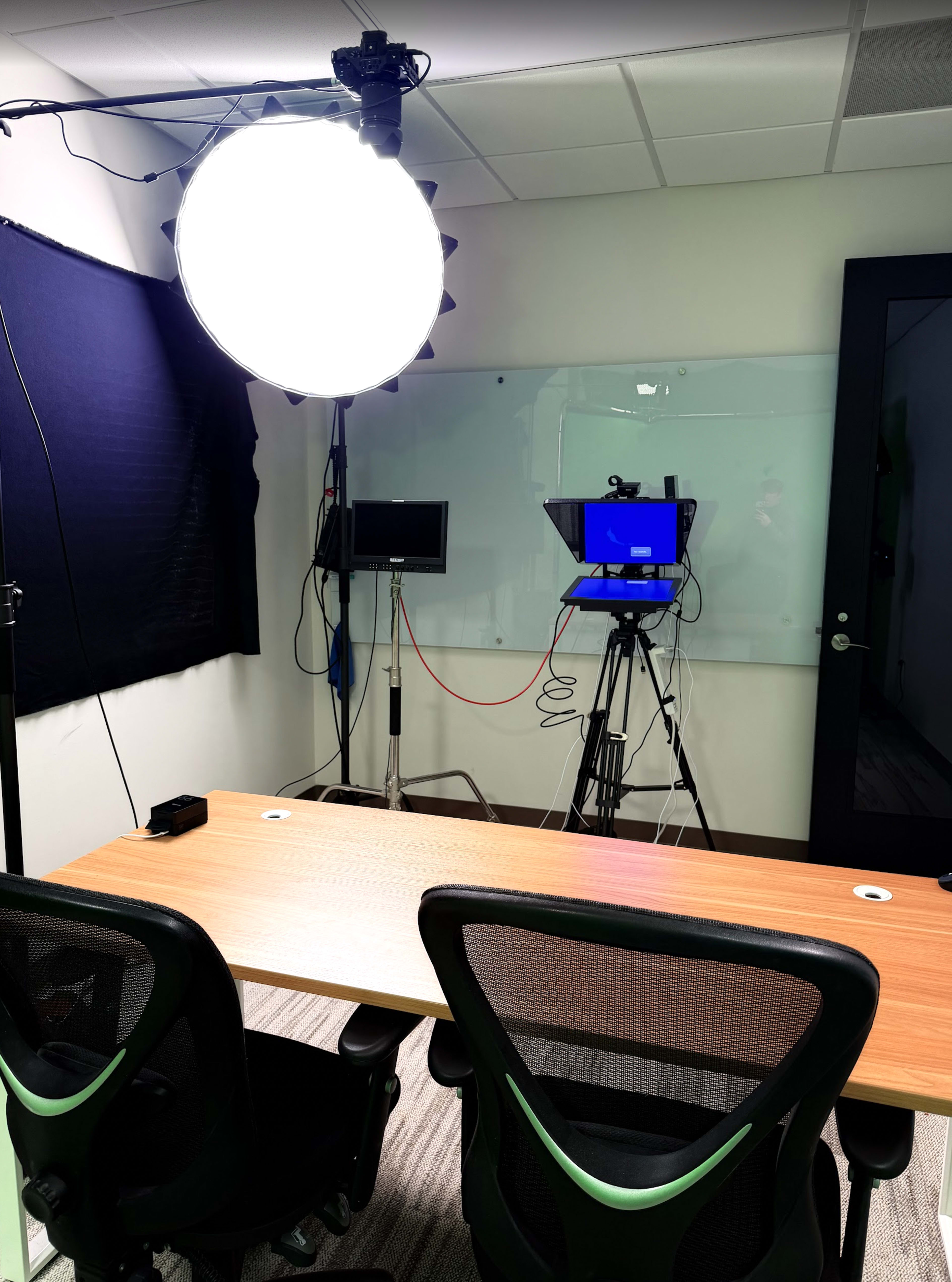 The image shows a well-lit room with a large circular light, a camera setup on a tripod, and a wooden table with two black office chairs.