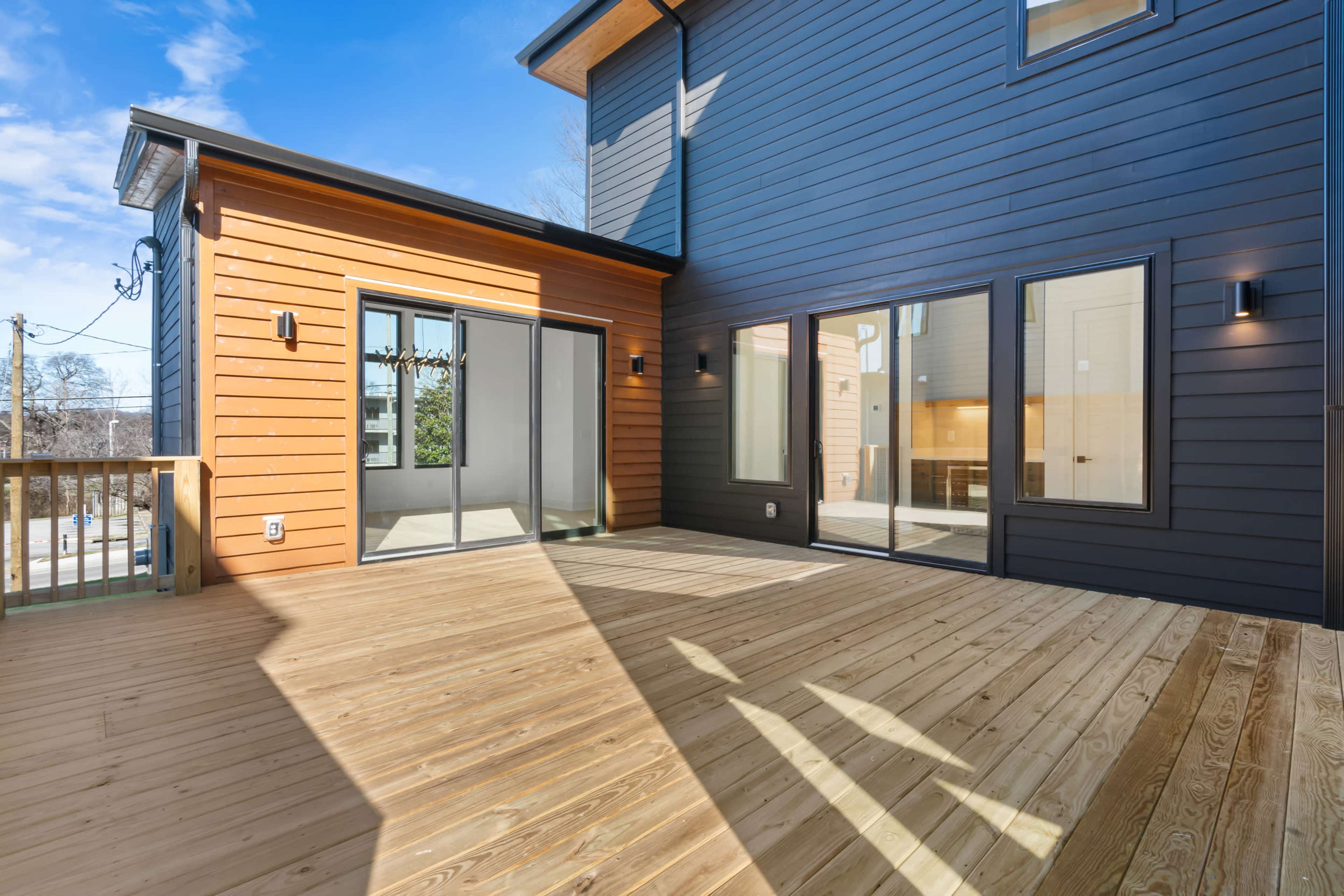 The image shows a modern wooden deck attached to a house with two-tone exterior siding and large glass doors leading indoors.