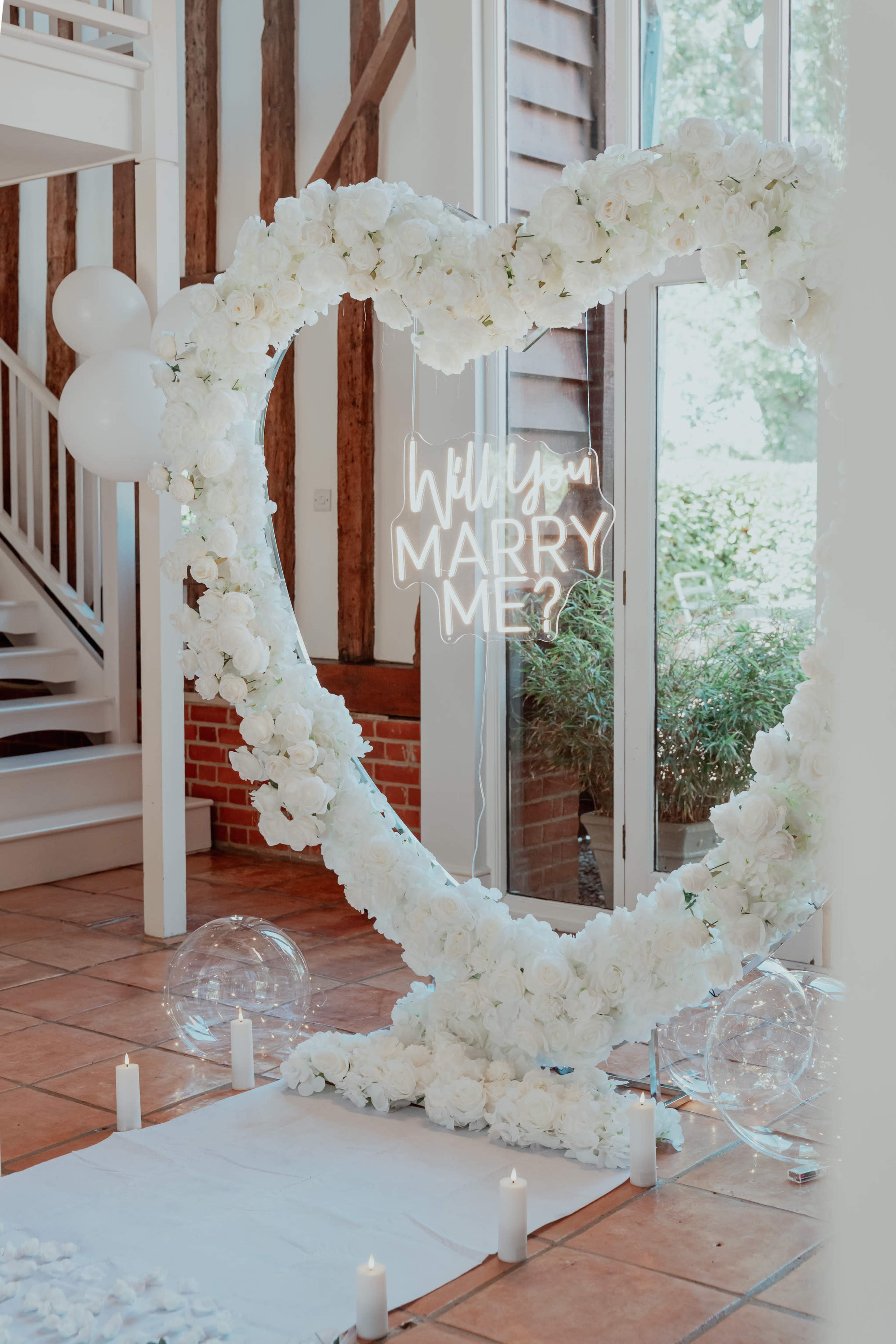 A heart-shaped floral arch with a neon sign reading "Will you marry me?" is set up indoors, surrounded by candles and transparent spheres.