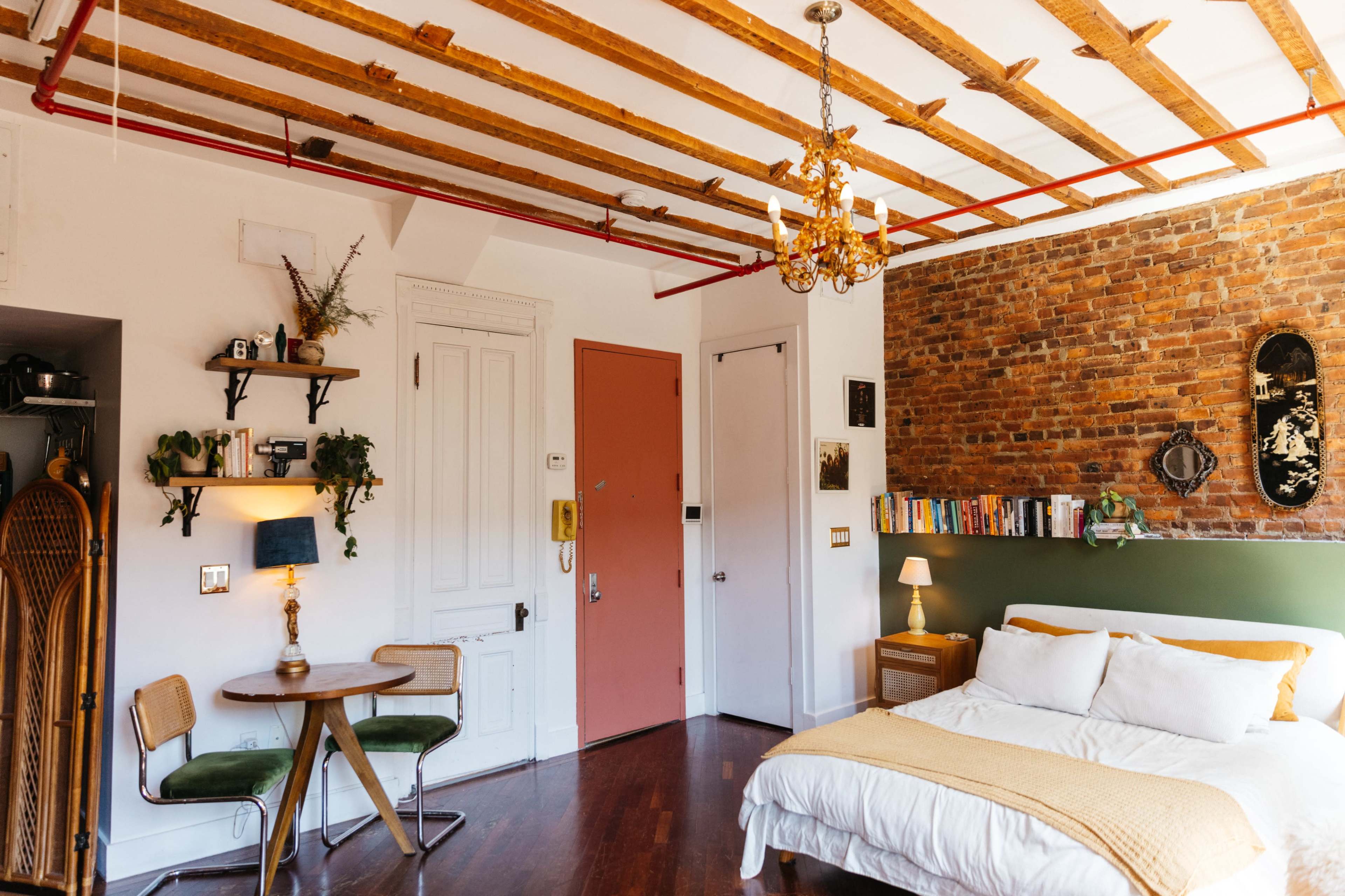 A cozy bedroom with exposed brick walls, a bed with a yellow bedspread, a small dining table, and various decorative shelves.