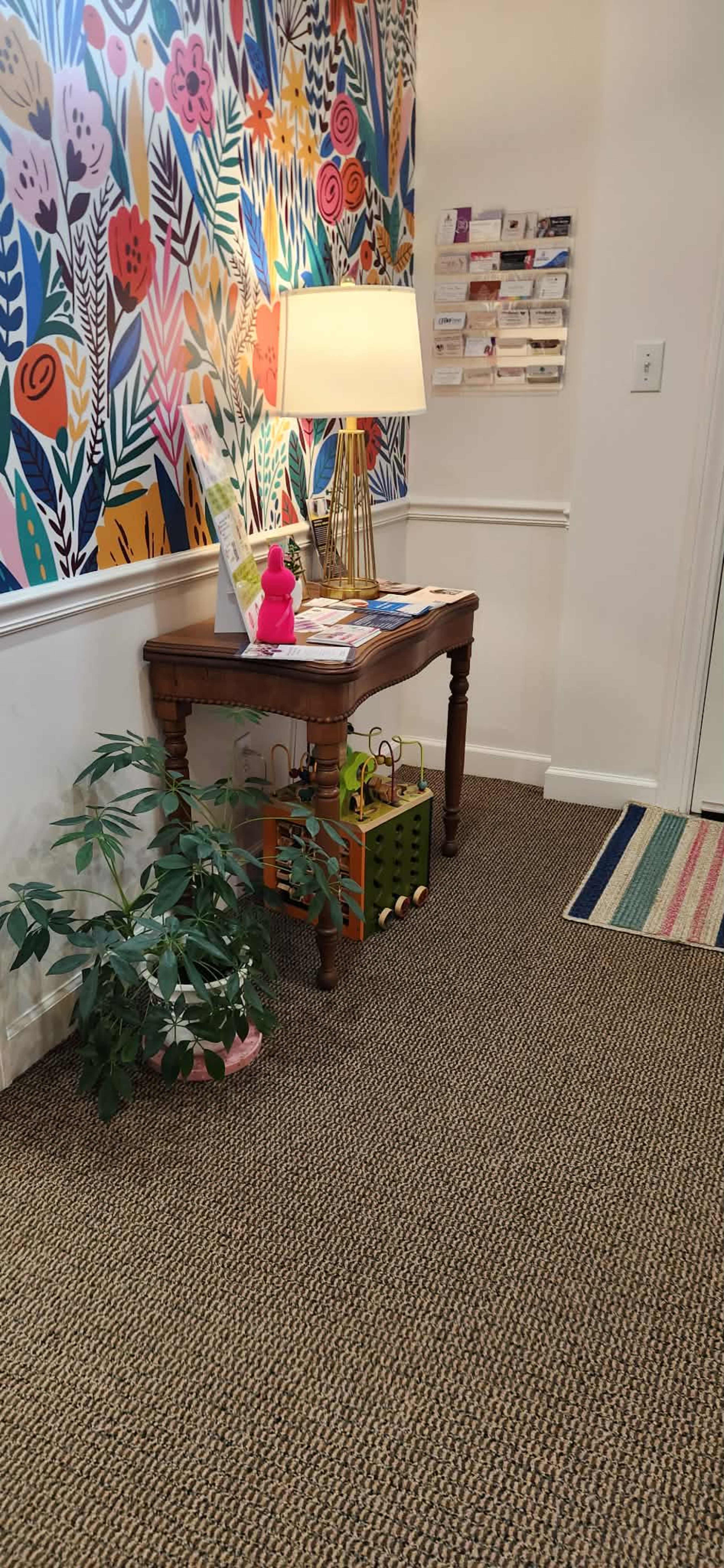The image shows a corner of a room featuring a wooden table with brochures, a lamp, and a colorful floral wallpaper; a green plant and a storage basket are on the floor beside the table.