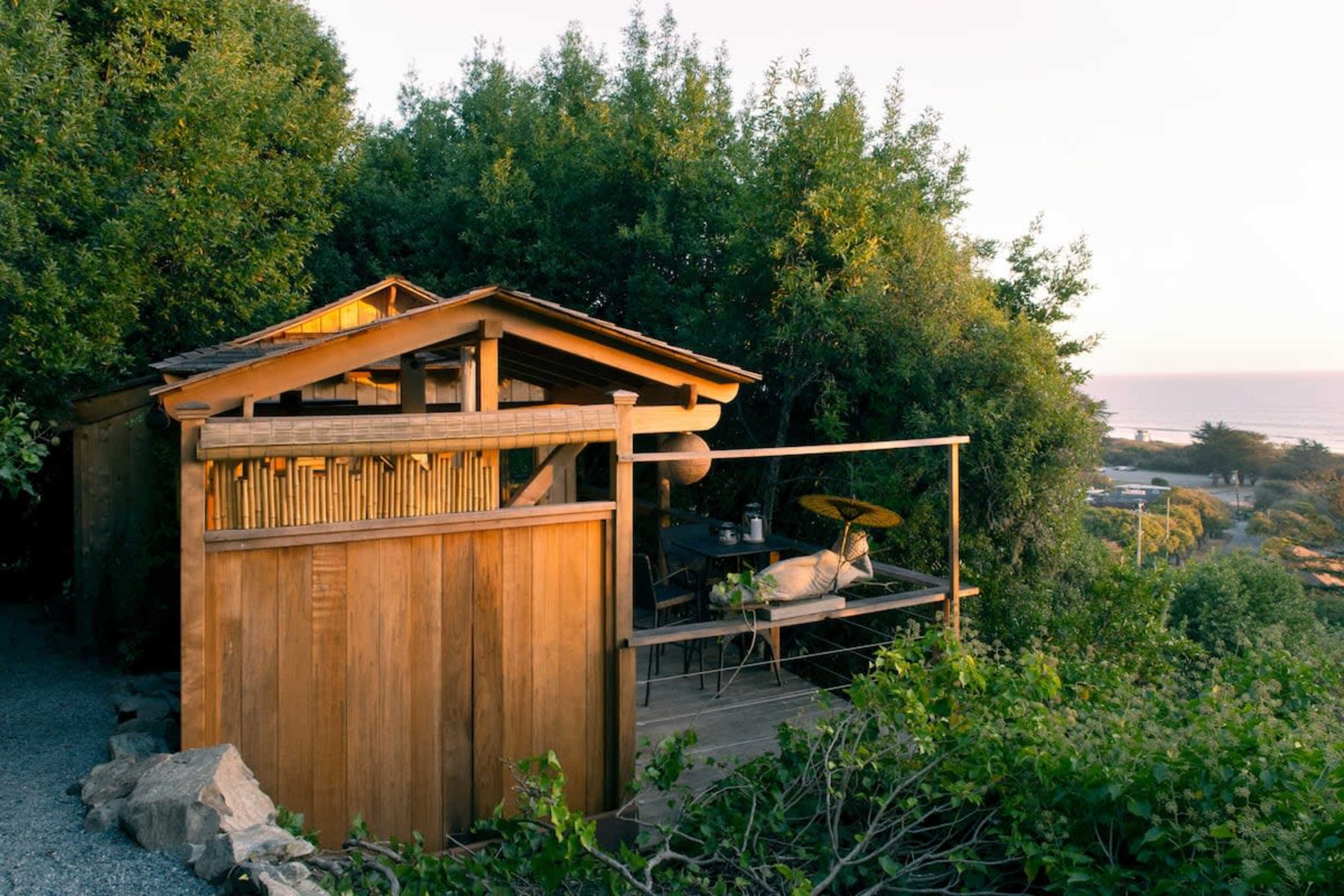A small wooden cabin is situated among trees, with a deck overlooking a coastal landscape.