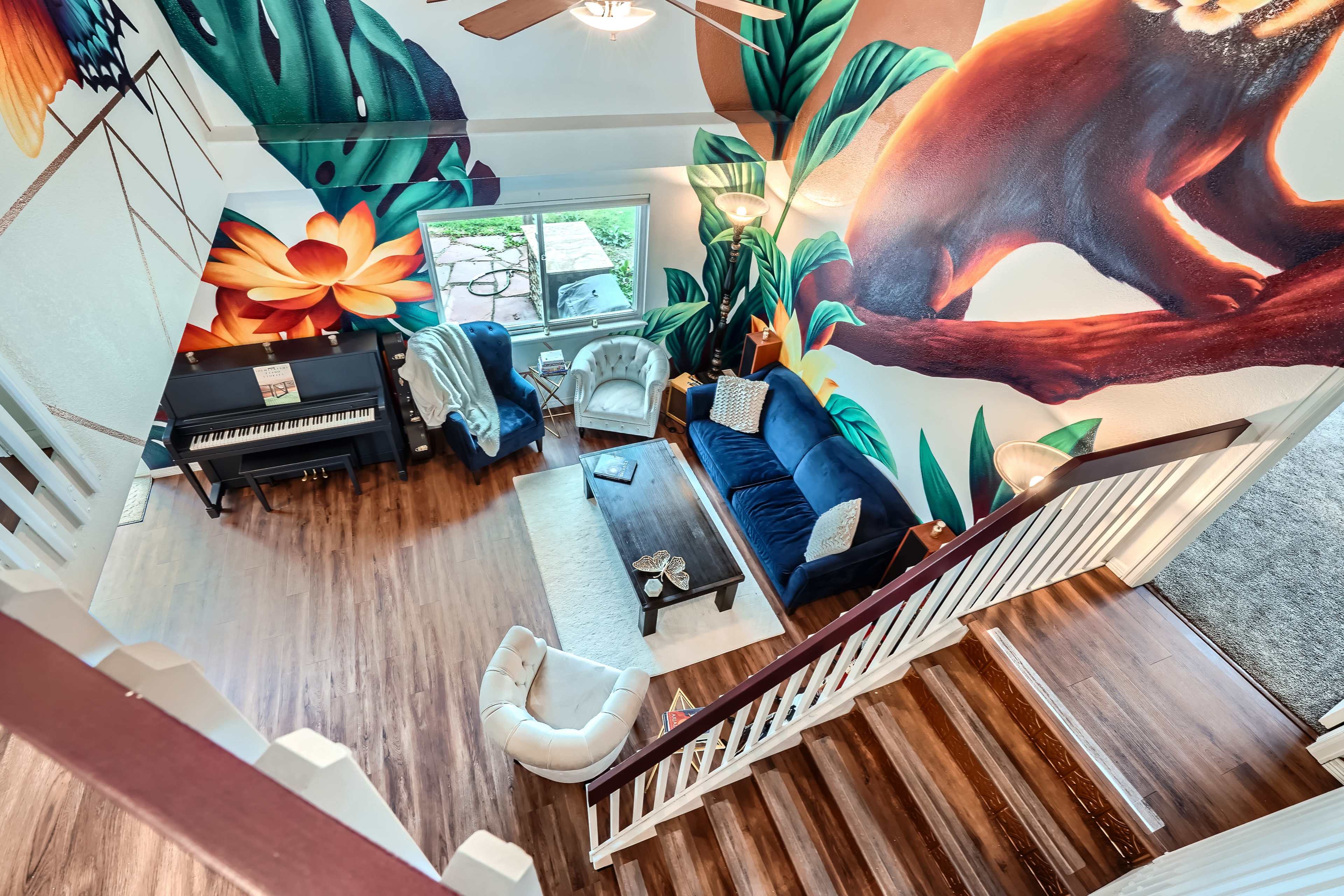 A living room with vibrant wall murals, a piano, a blue sofa, and a staircase leading to another area.
