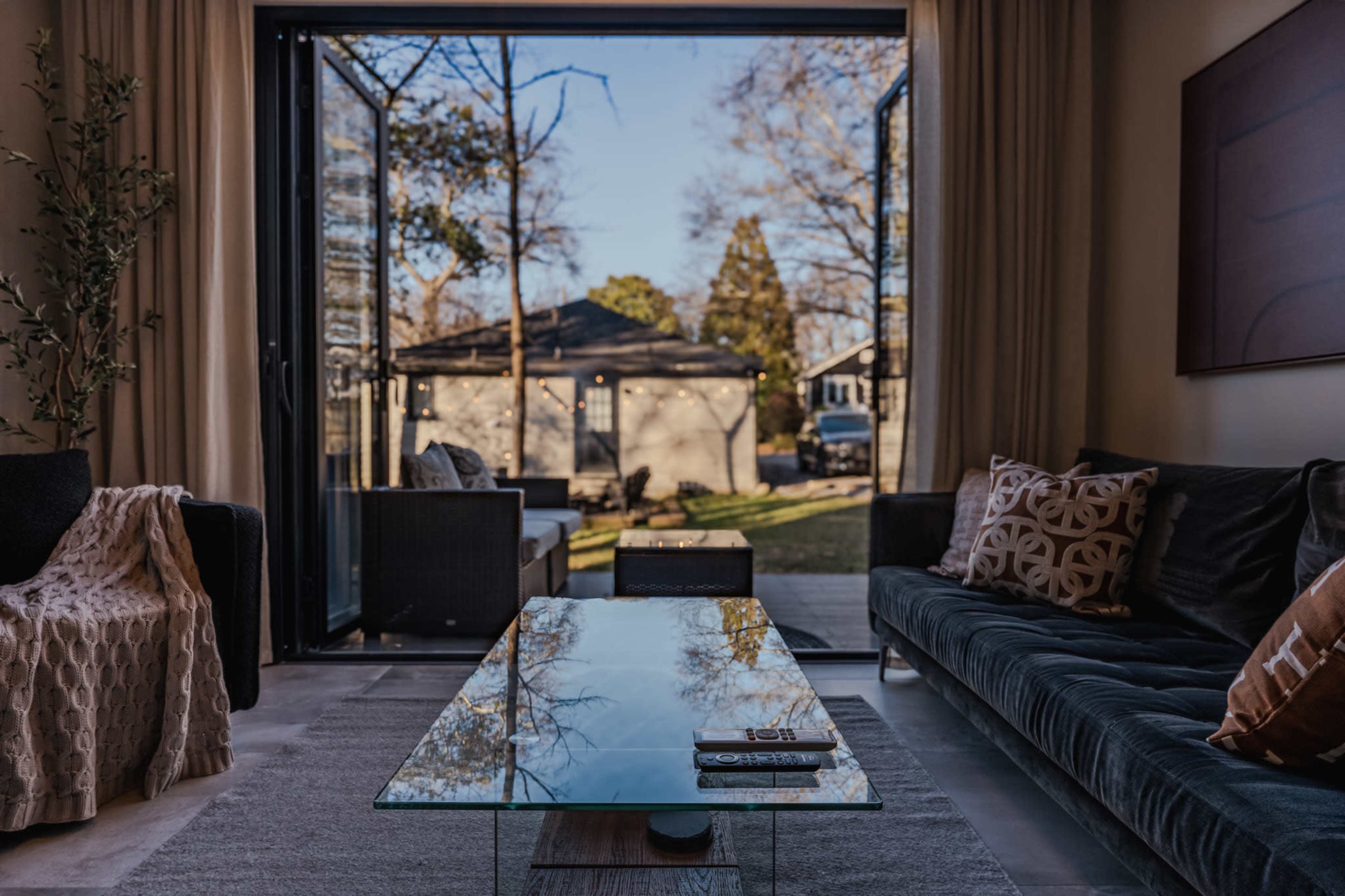A modern living room features a glass coffee table and seating, with large sliding doors open to a view of an outdoor patio and garden.