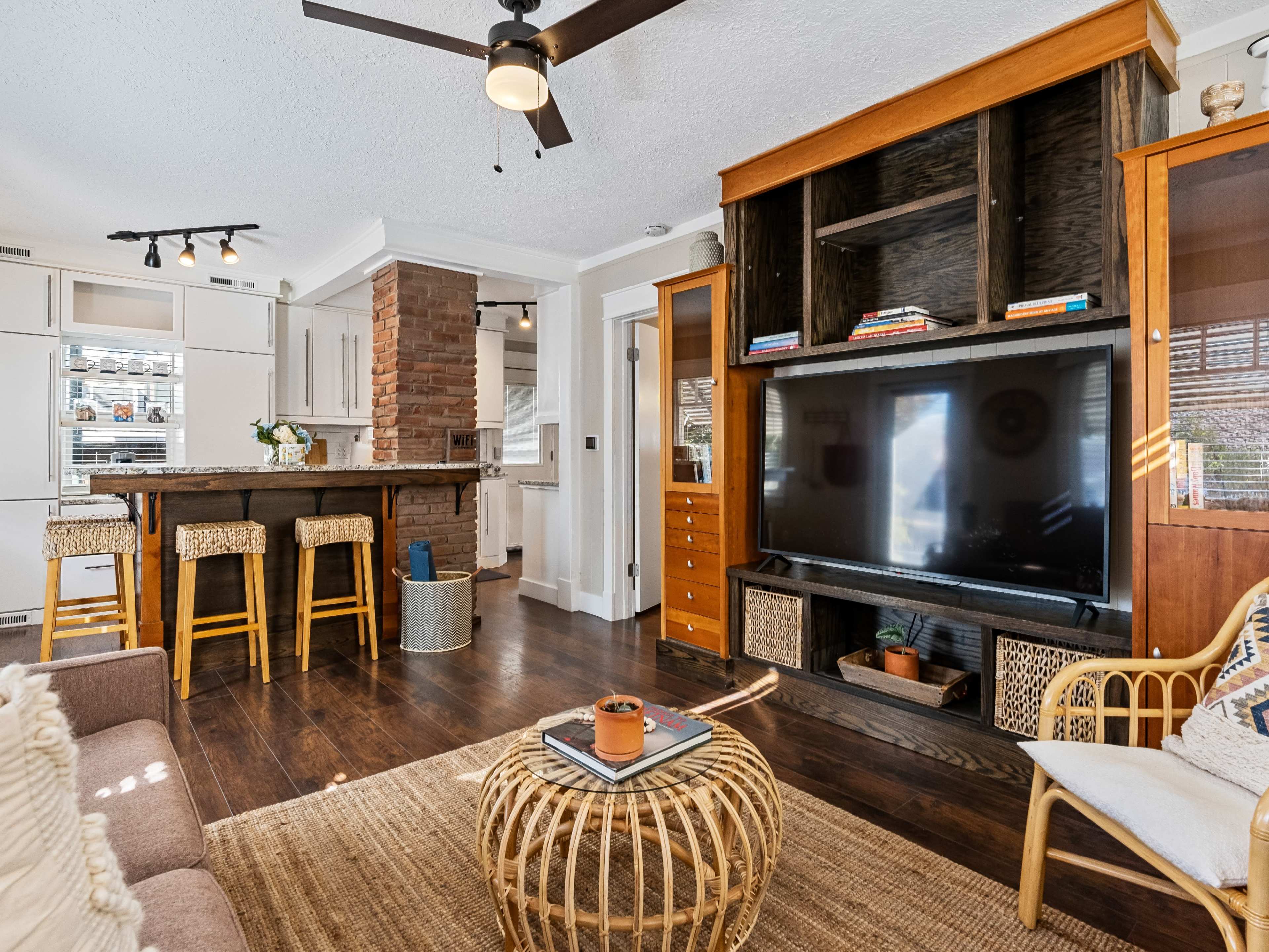 A modern living room featuring a television unit, a cozy seating area, and a kitchen bar with stools in the background.