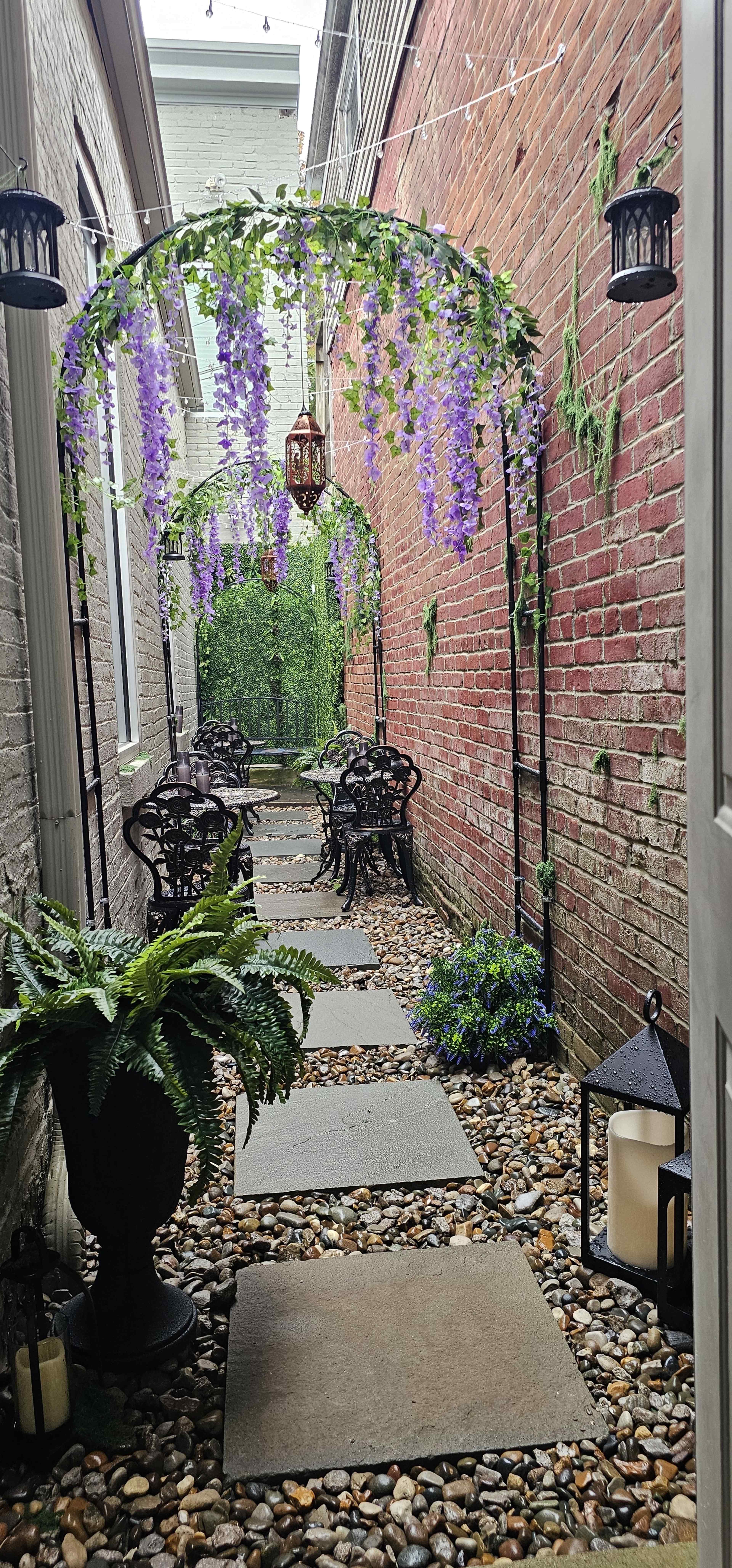 The image shows a narrow outdoor pathway lined with stone slabs, surrounded by brick walls and adorned with hanging purple flowers, lanterns, and greenery.