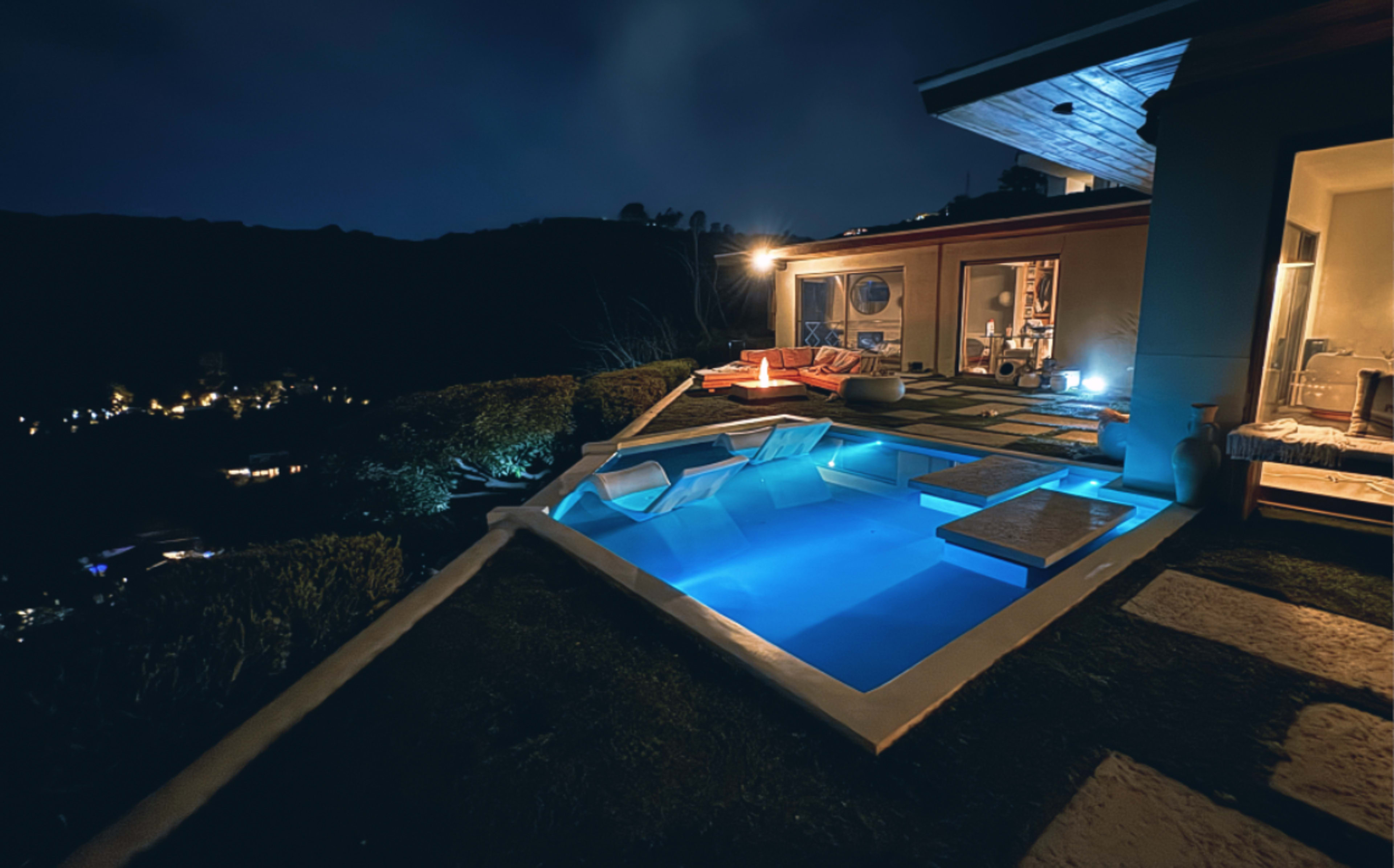 A modern pool with illuminated water is situated on a hillside terrace, overlooking a landscape of distant lights at night.