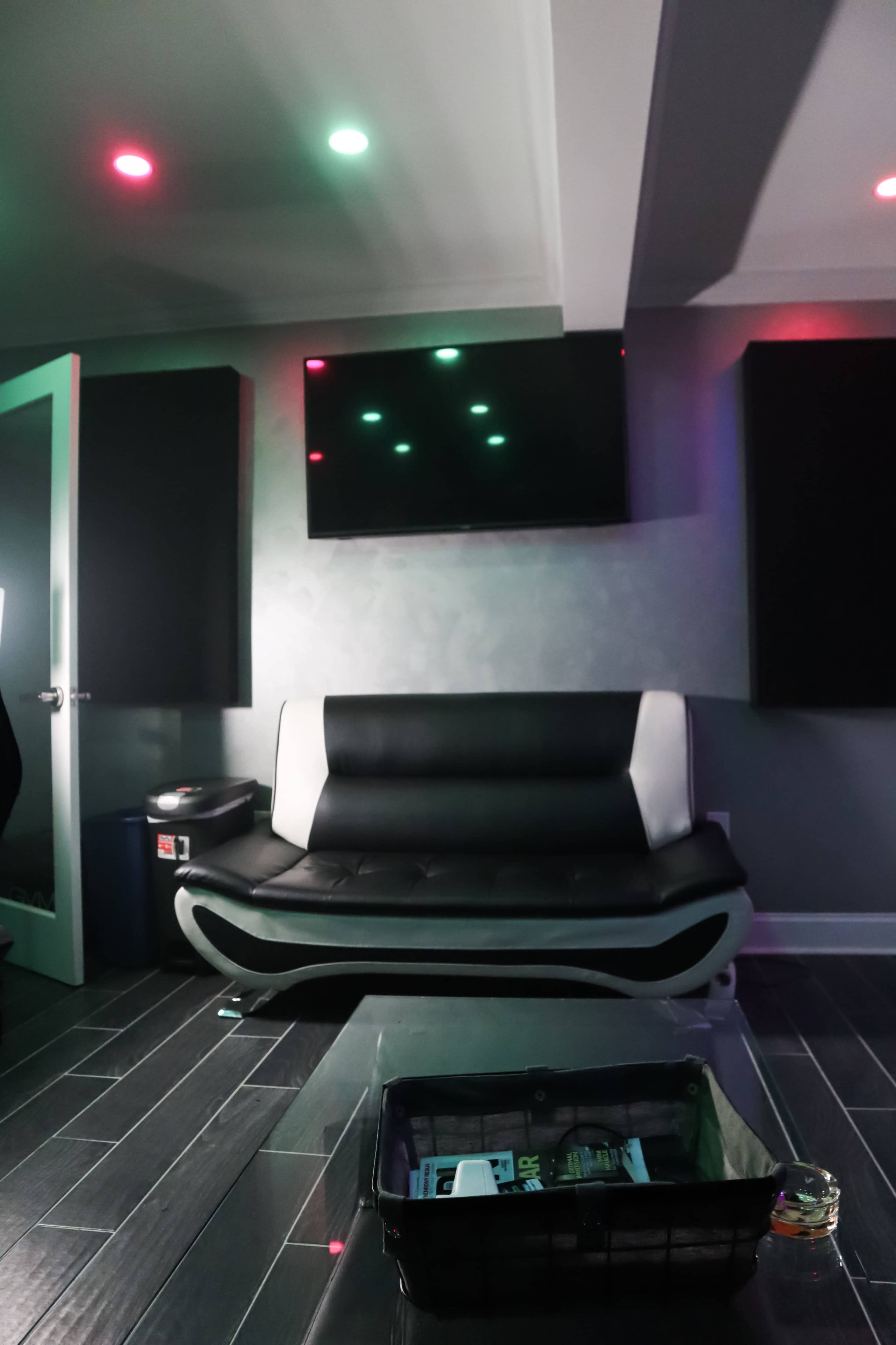 The image shows a modern lounge area with a black and white sofa, a flat-screen TV mounted on the wall, and soft colored lights illuminating the space.