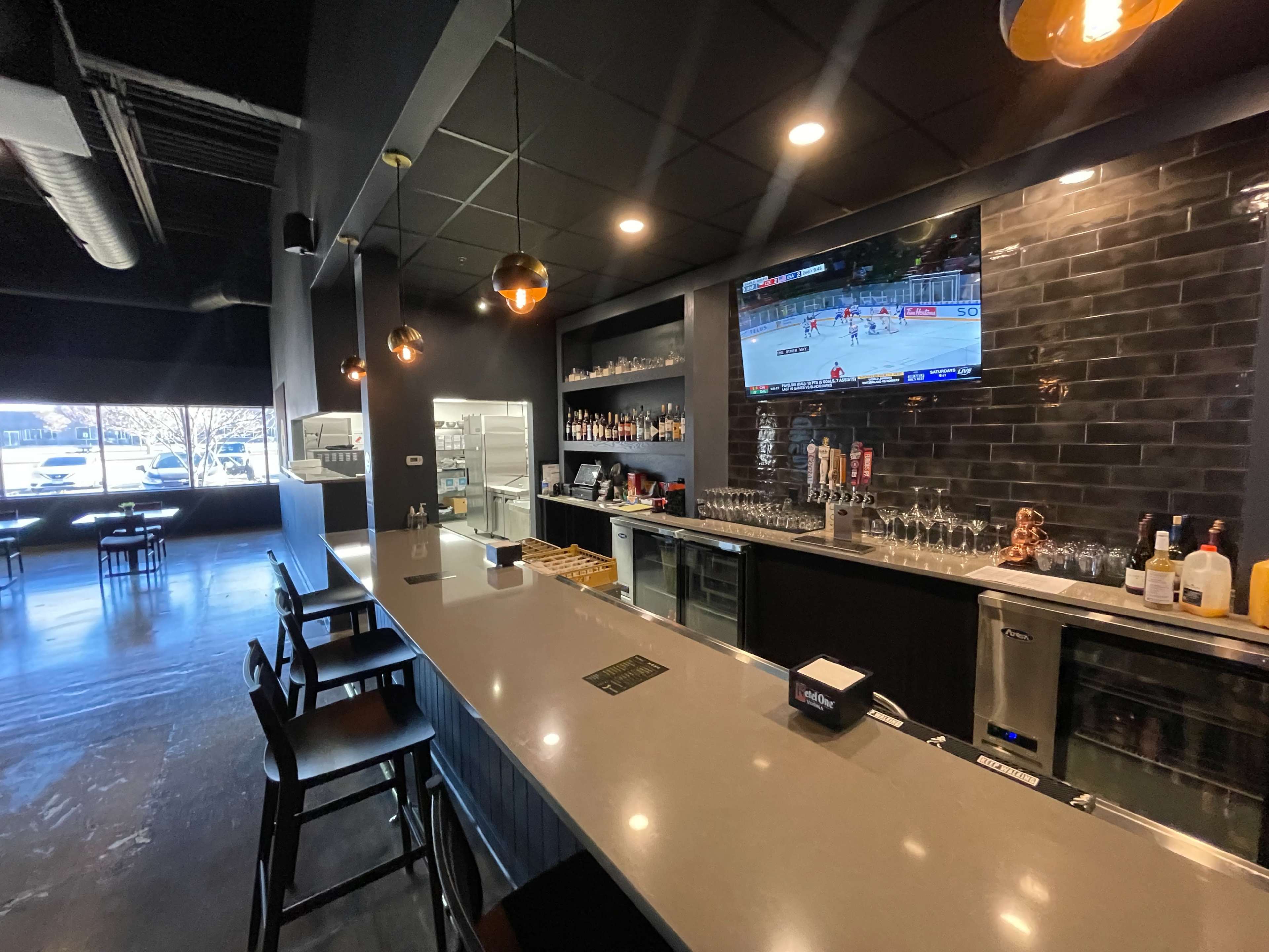 The image shows a modern bar area with a counter, stools, a television displaying a sports game, and a well-stocked shelf behind the bar.