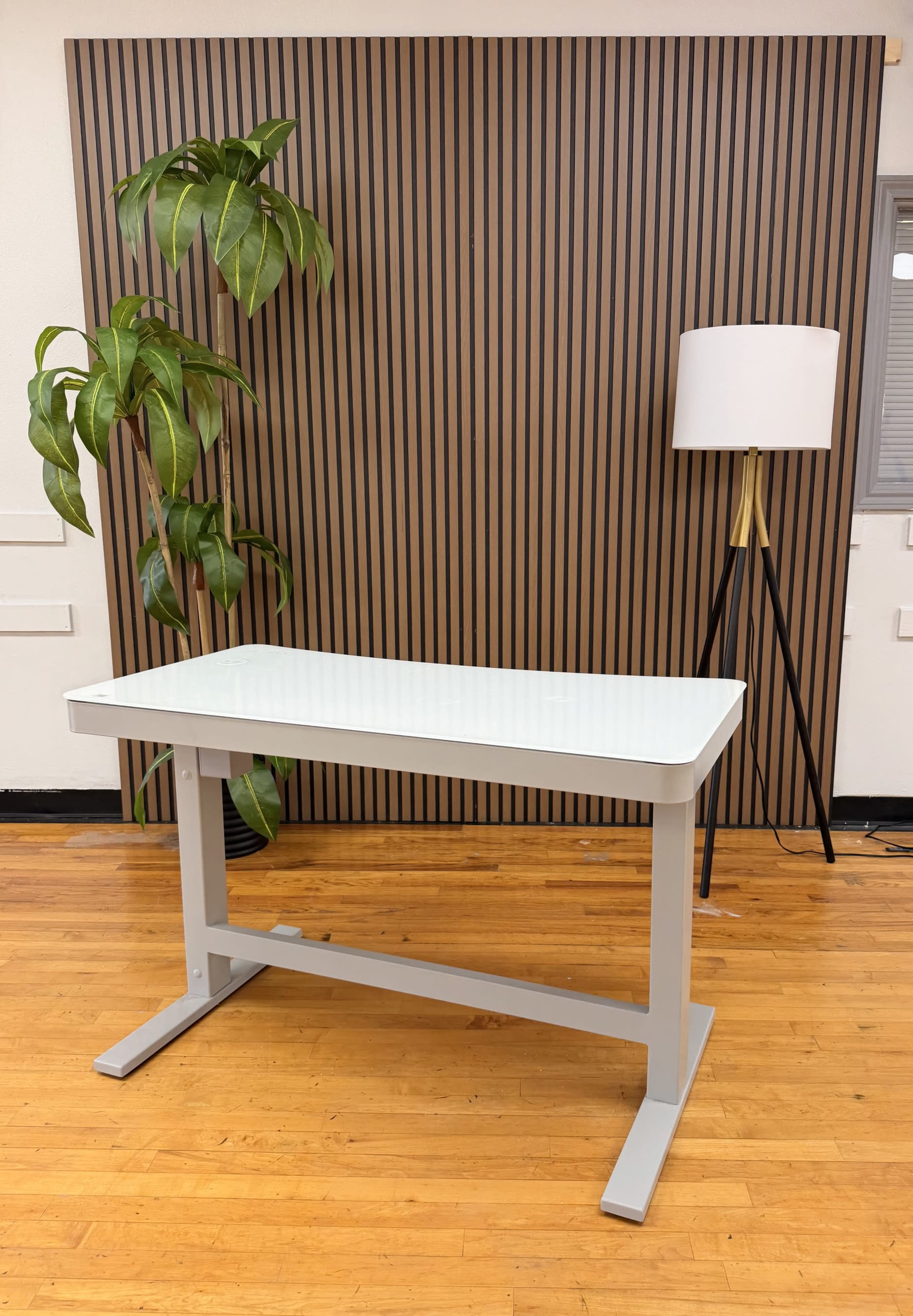 The image shows a modern, minimalist desk with a sleek white surface set against a striped wooden wall, accompanied by a potted plant and a tall lamp nearby.