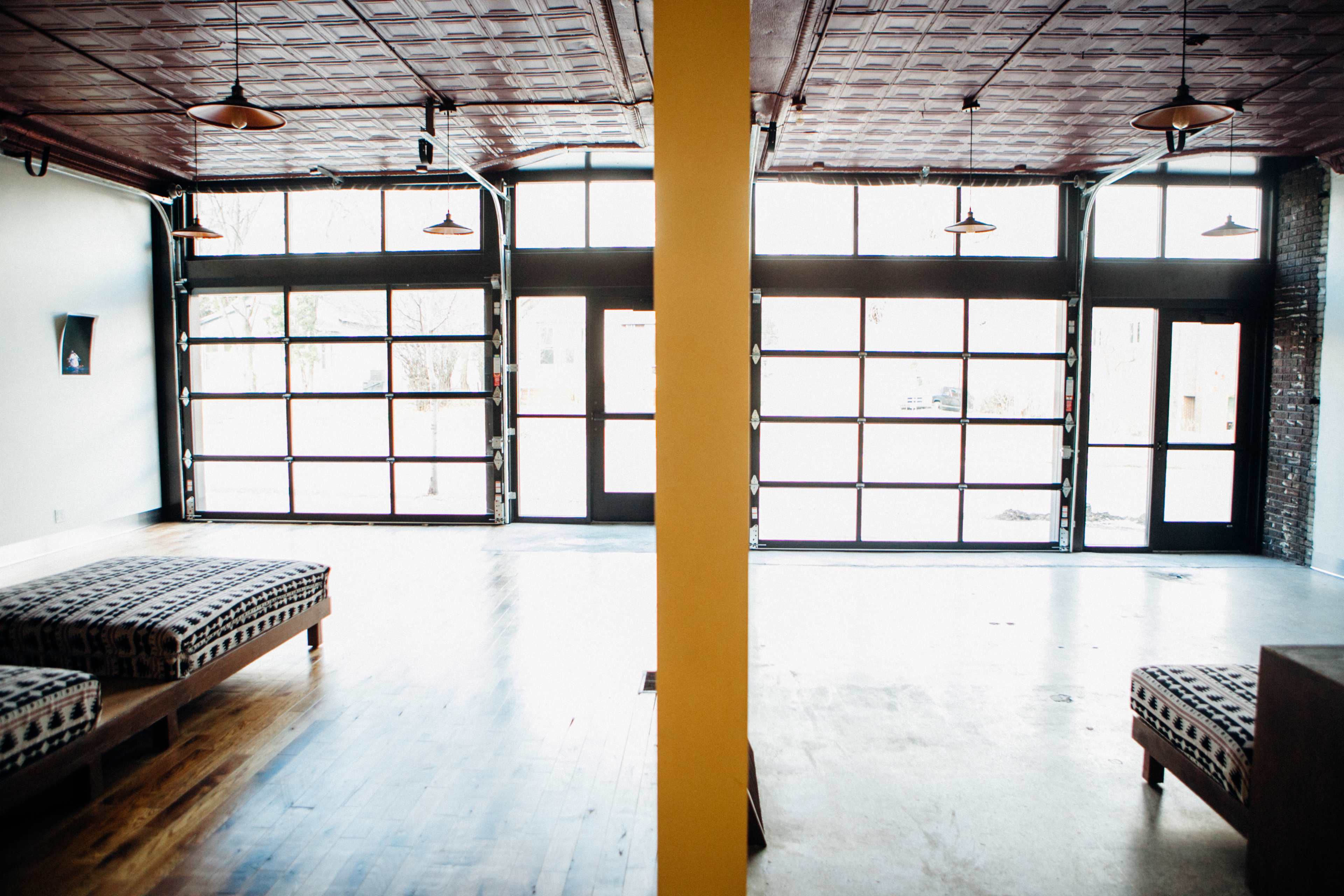 The image shows an empty interior space with polished wooden floors, a combination of large glass doors and windows, and two seating areas divided by a yellow column.