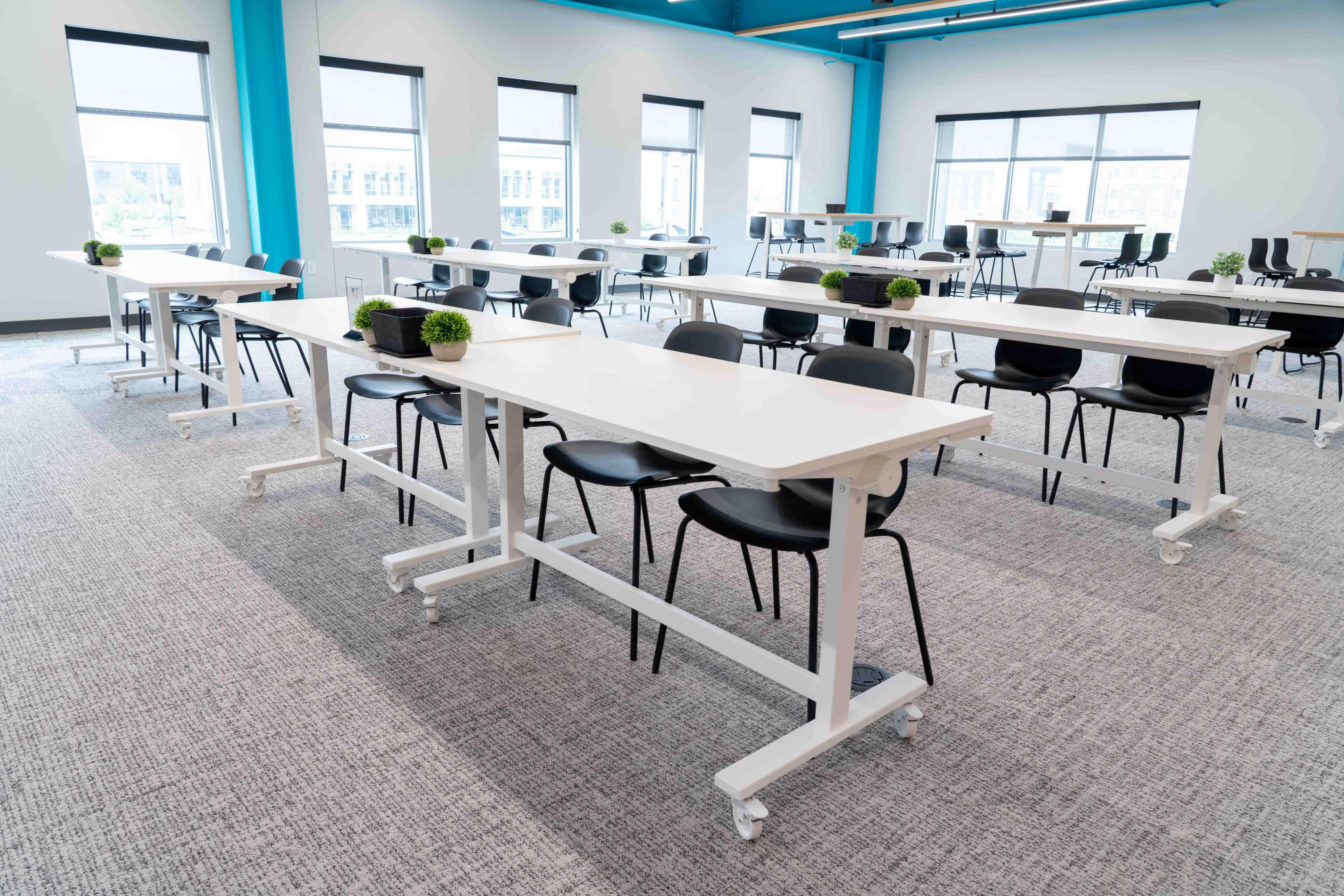 The image shows a modern, open room with several white tables on wheels, each accompanied by black chairs and small potted plants.