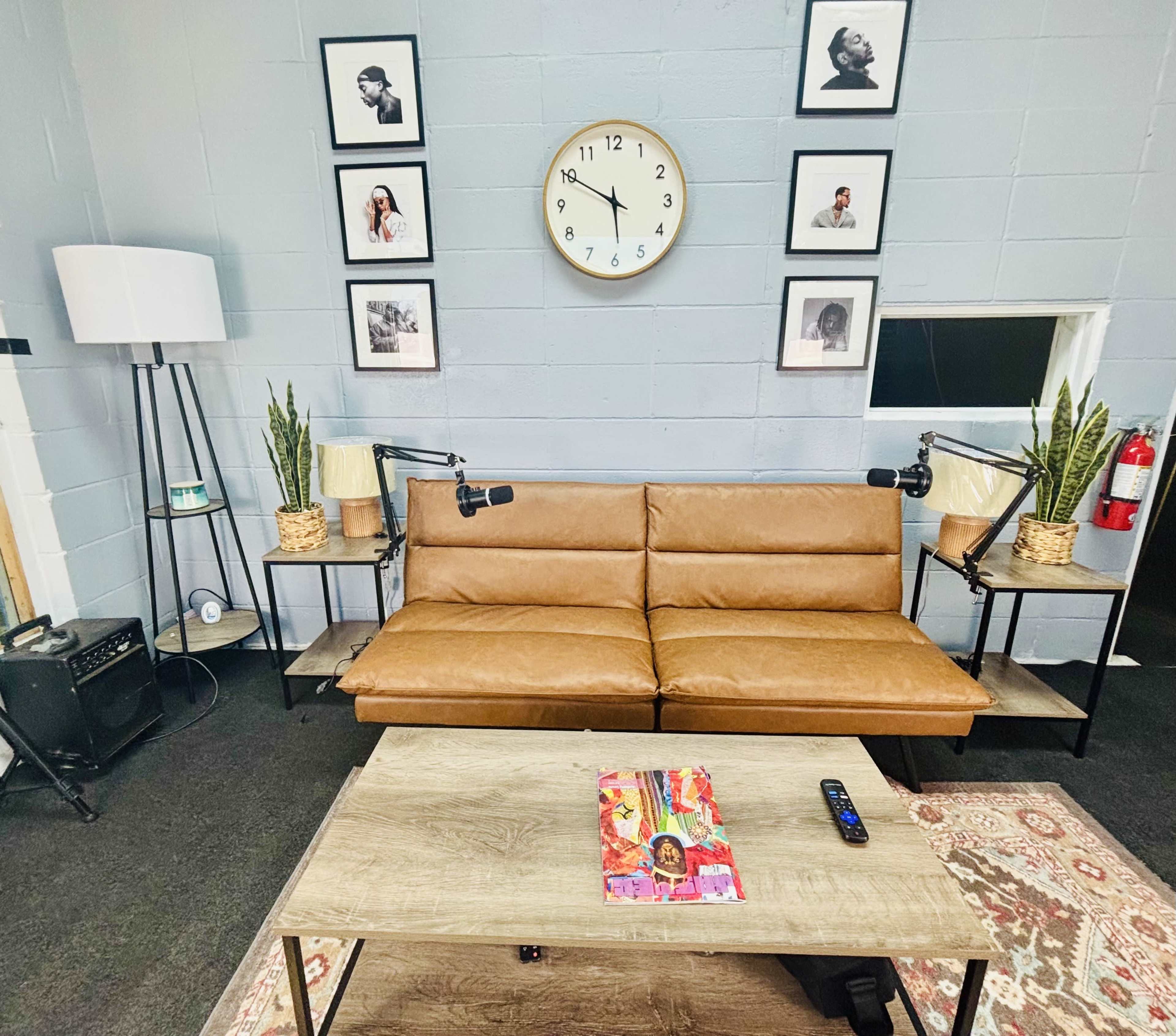 The image shows a cozy recording studio with a brown leather couch, a wooden coffee table, and framed black-and-white photos on the wall, accompanied by two microphones and decorative plants.