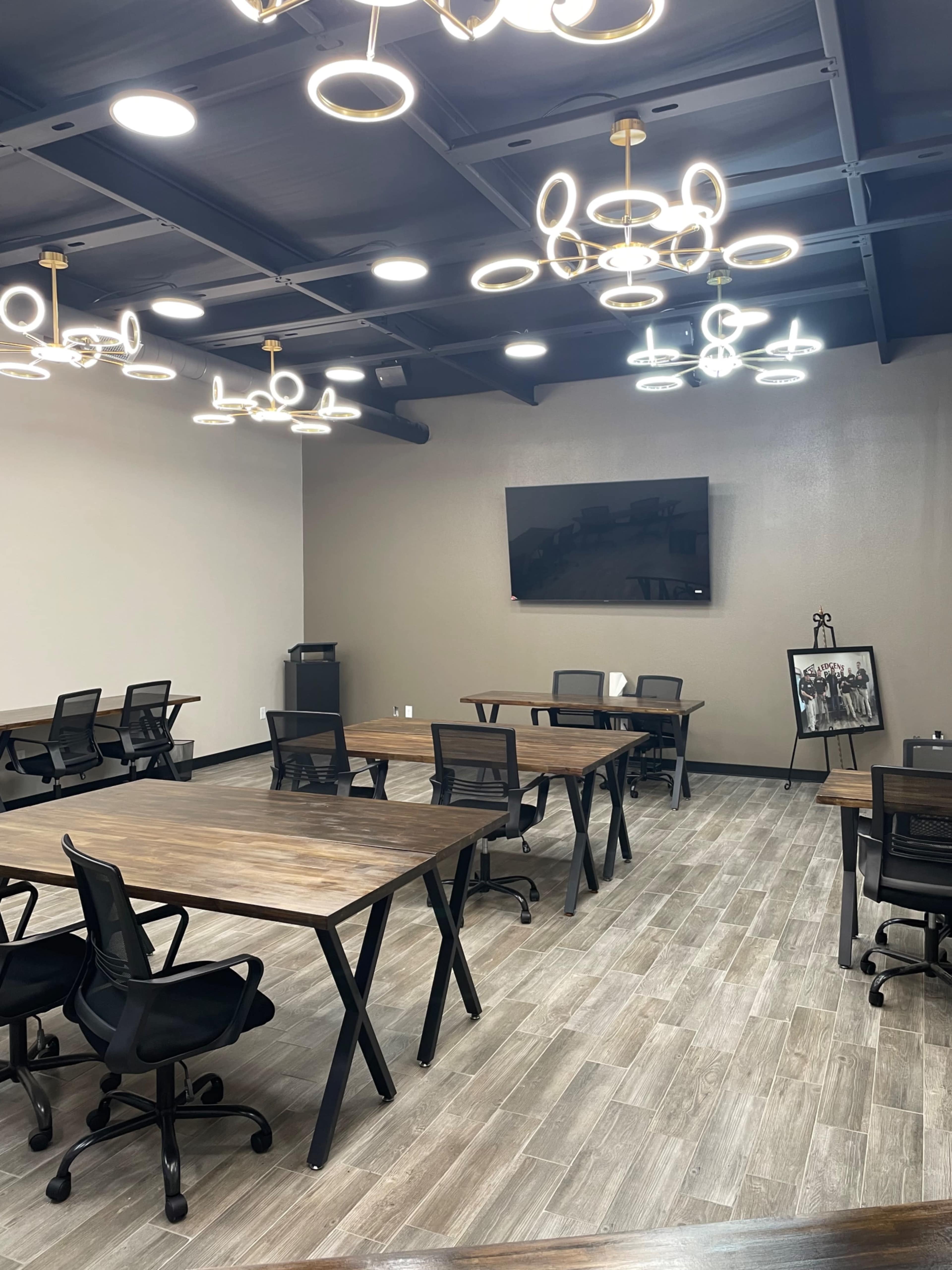 A modern meeting room with several wooden tables, black chairs, and multiple light fixtures hanging from the ceiling.