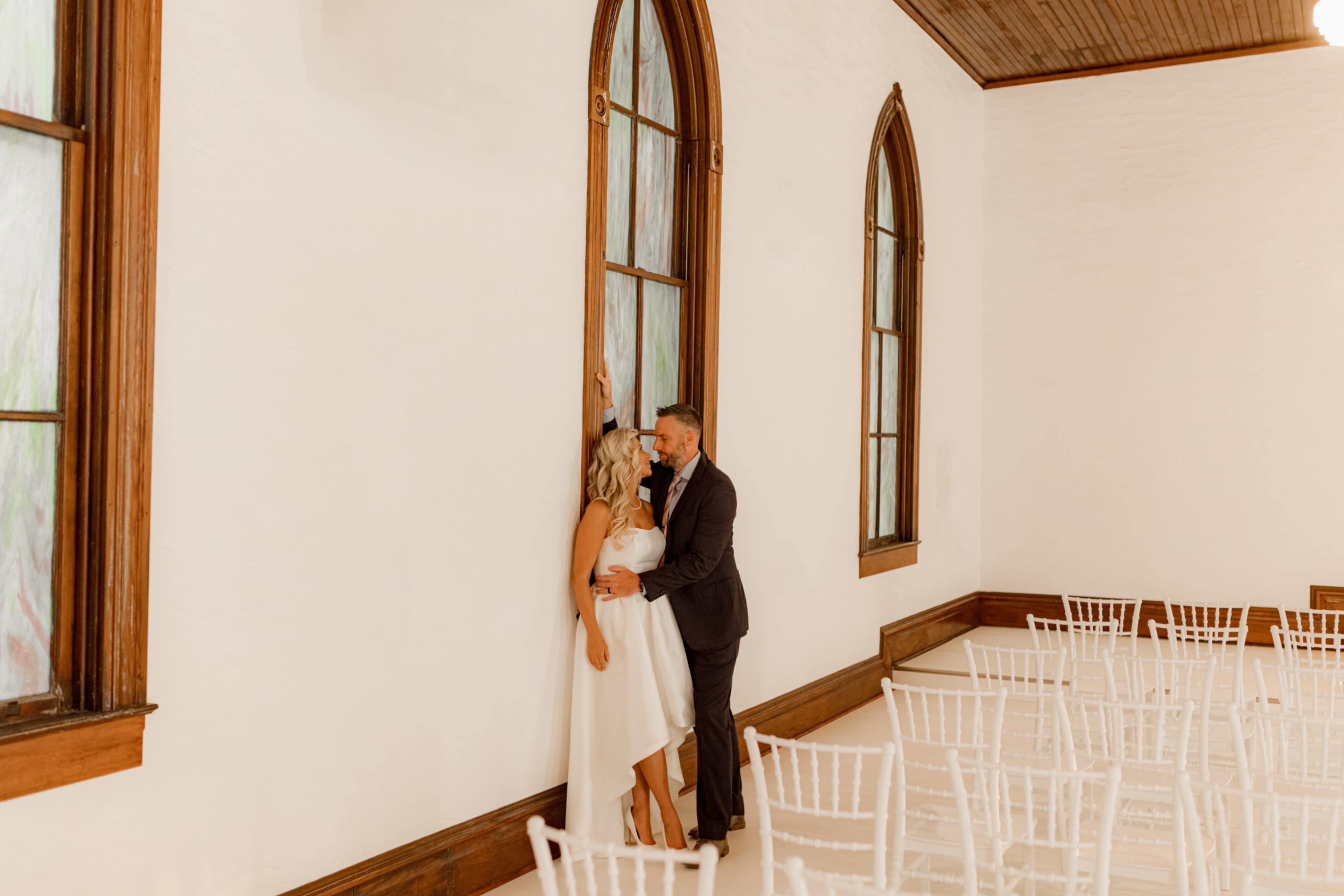 A couple stands close together near a large window in a sparsely furnished room with white walls and wooden accents.