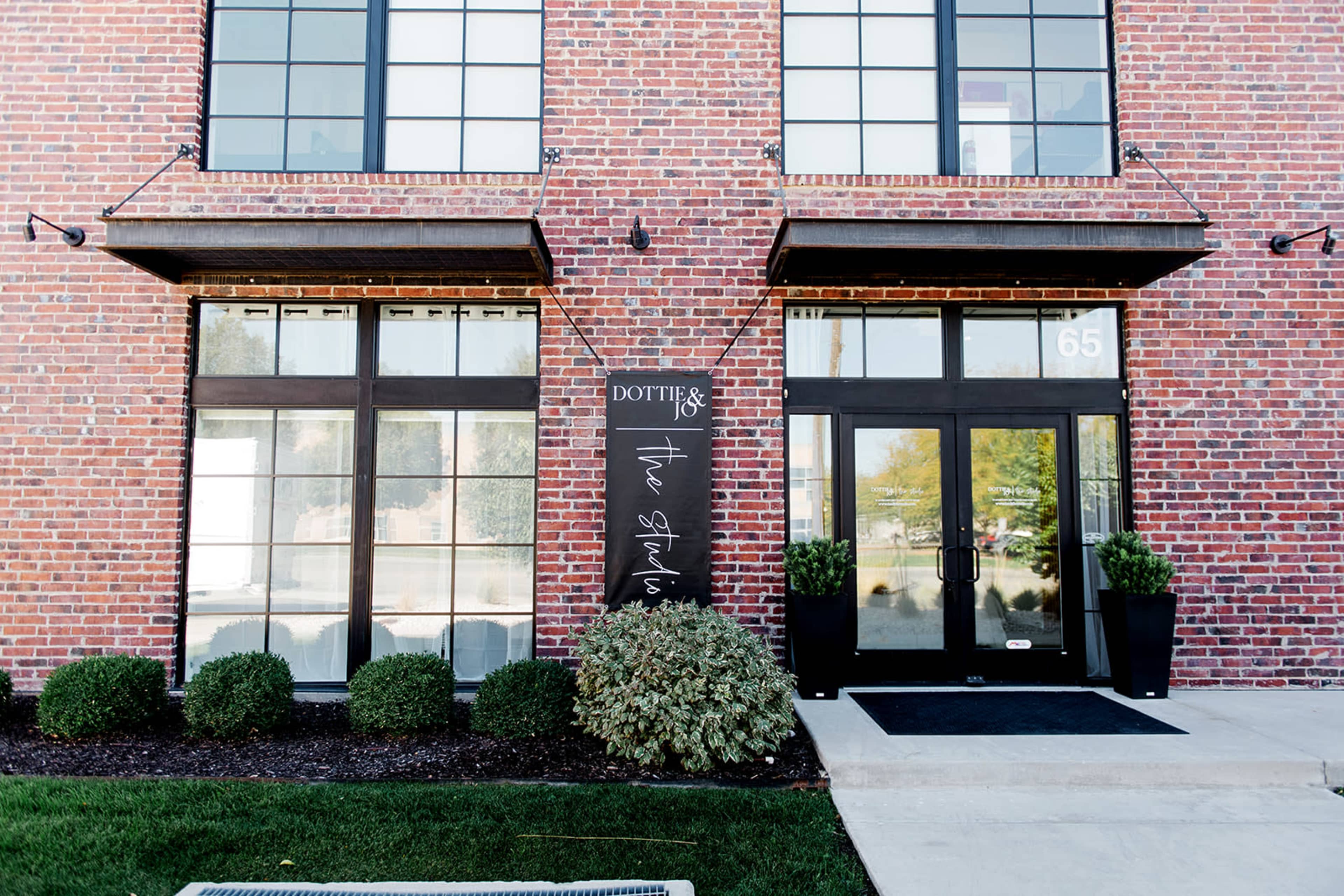 The image shows a brick building with large windows and two entrance doors, framed by neatly trimmed bushes and a sign labeled "The Studios."