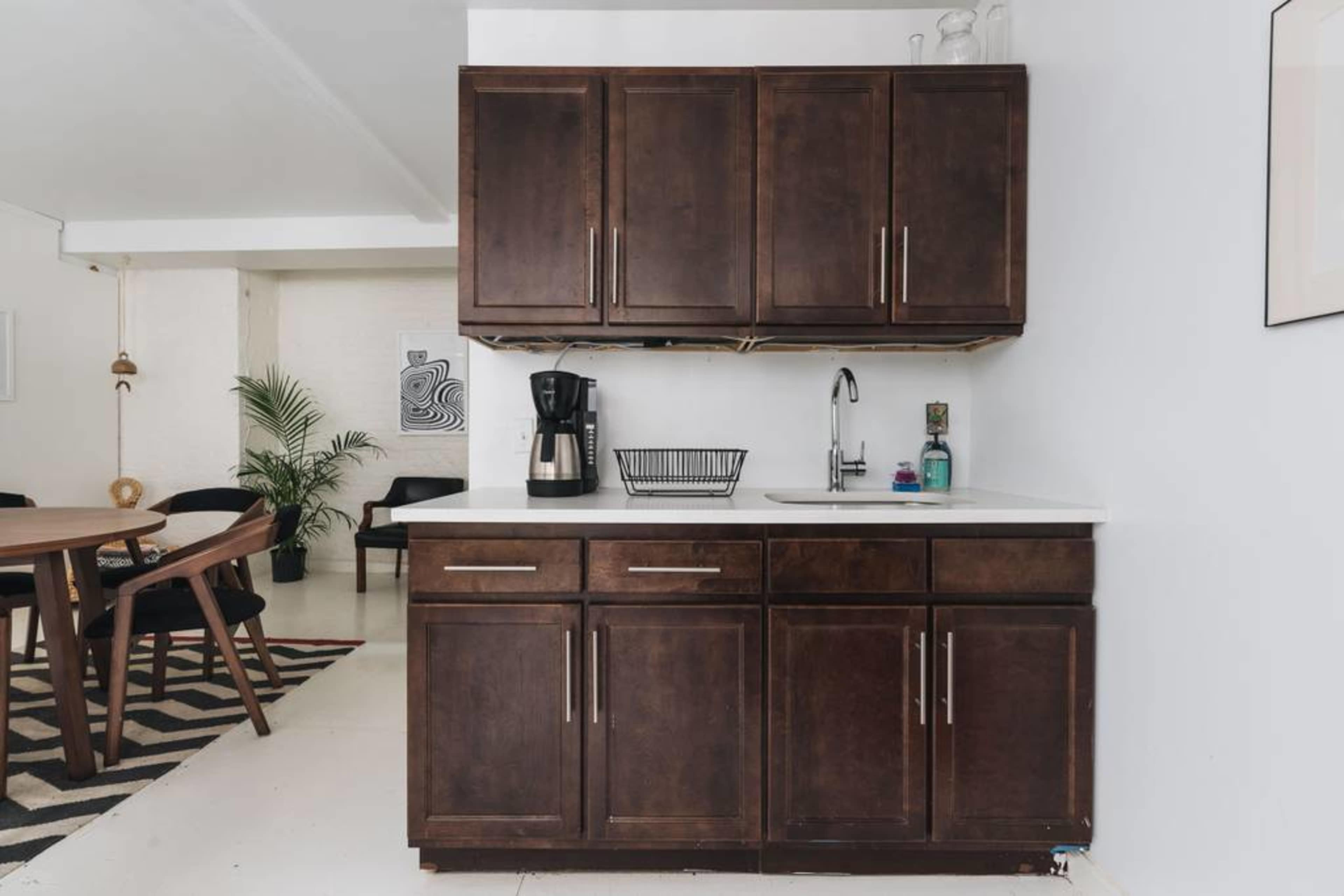 A kitchen area featuring dark wood cabinets, a coffee maker, a sink, and a dining area in the background with a patterned rug.
