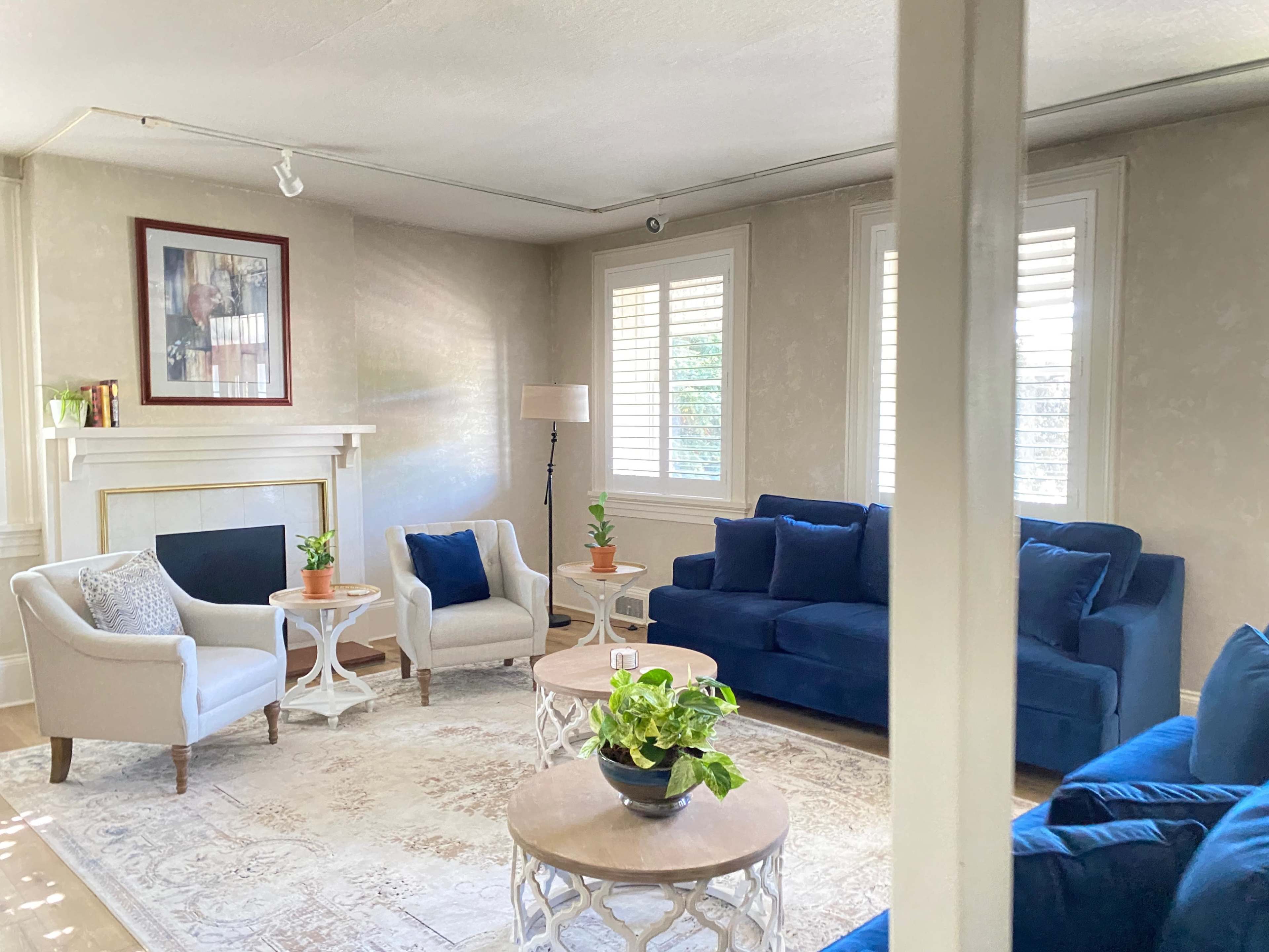 A living room with a blue sofa, two light-colored armchairs, a patterned rug, and potted plants on circular coffee tables.