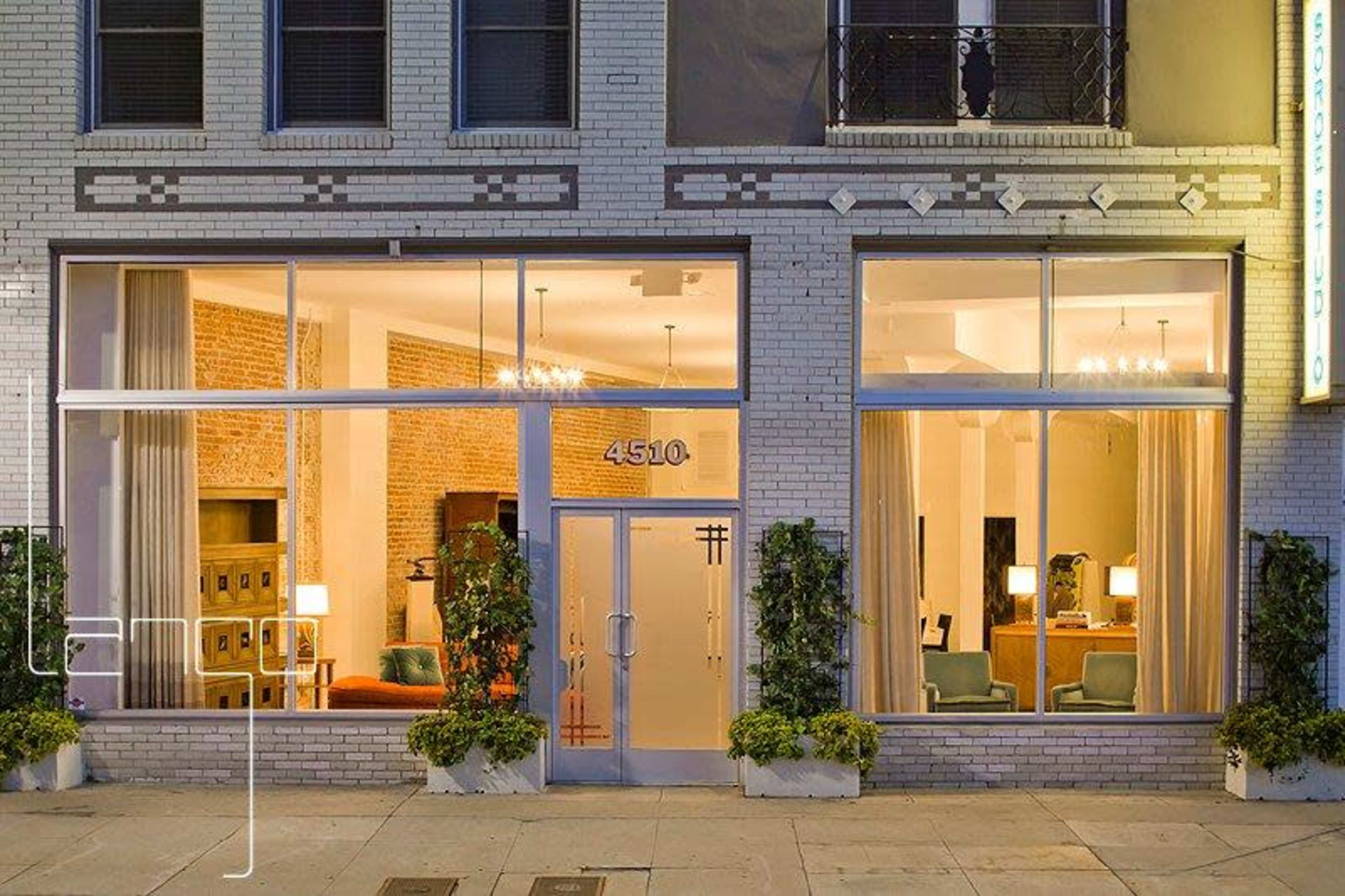 A modern building facade with large glass windows displaying an interior living space furnished with seating and lighting fixtures.