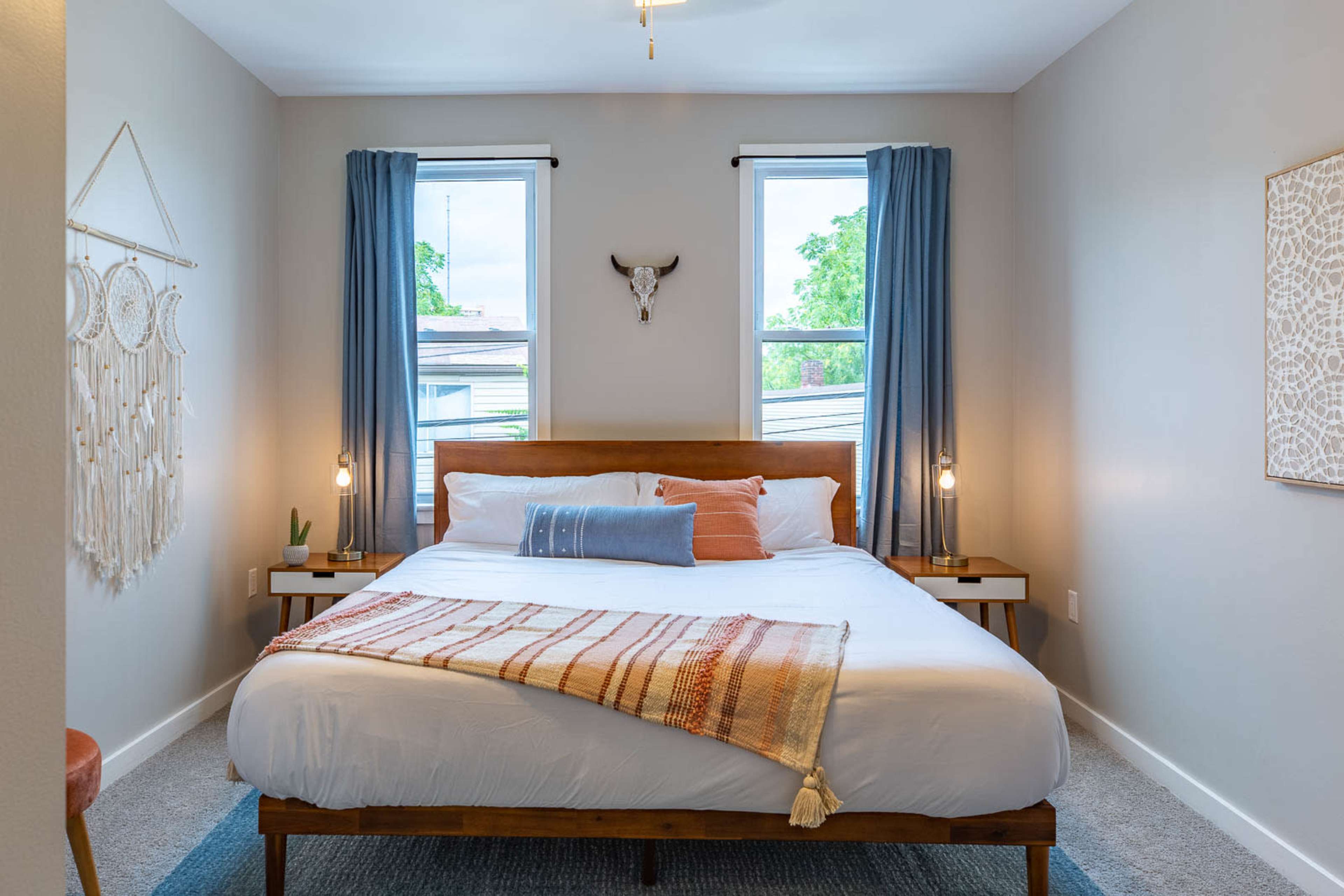 A neatly arranged bedroom features a wooden bed with white bedding, two bedside tables, and blue curtains framing the windows.