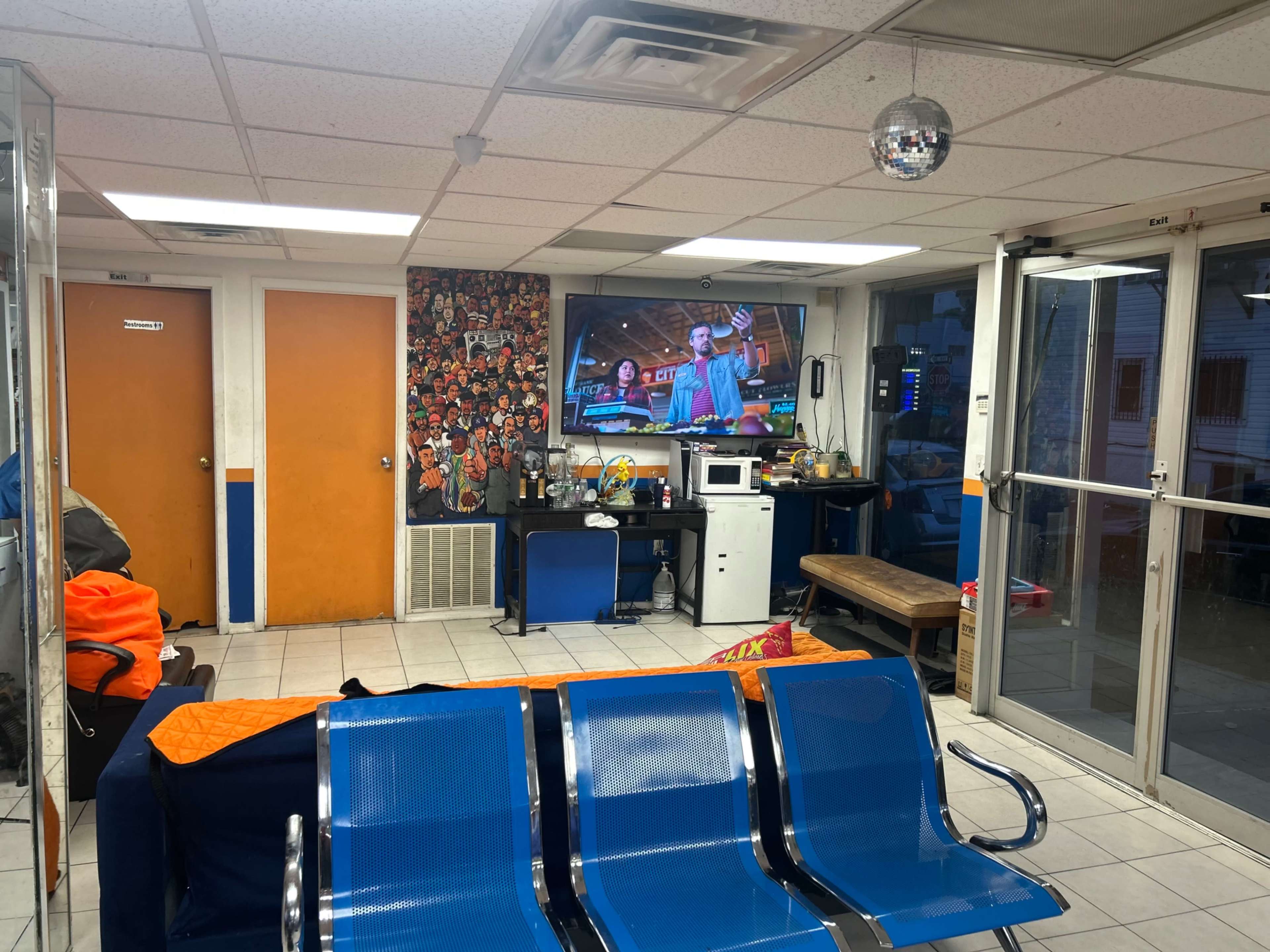 The image shows a waiting area with metal chairs, a television mounted on the wall, a microwave, and a small table, surrounded by orange and blue decor.