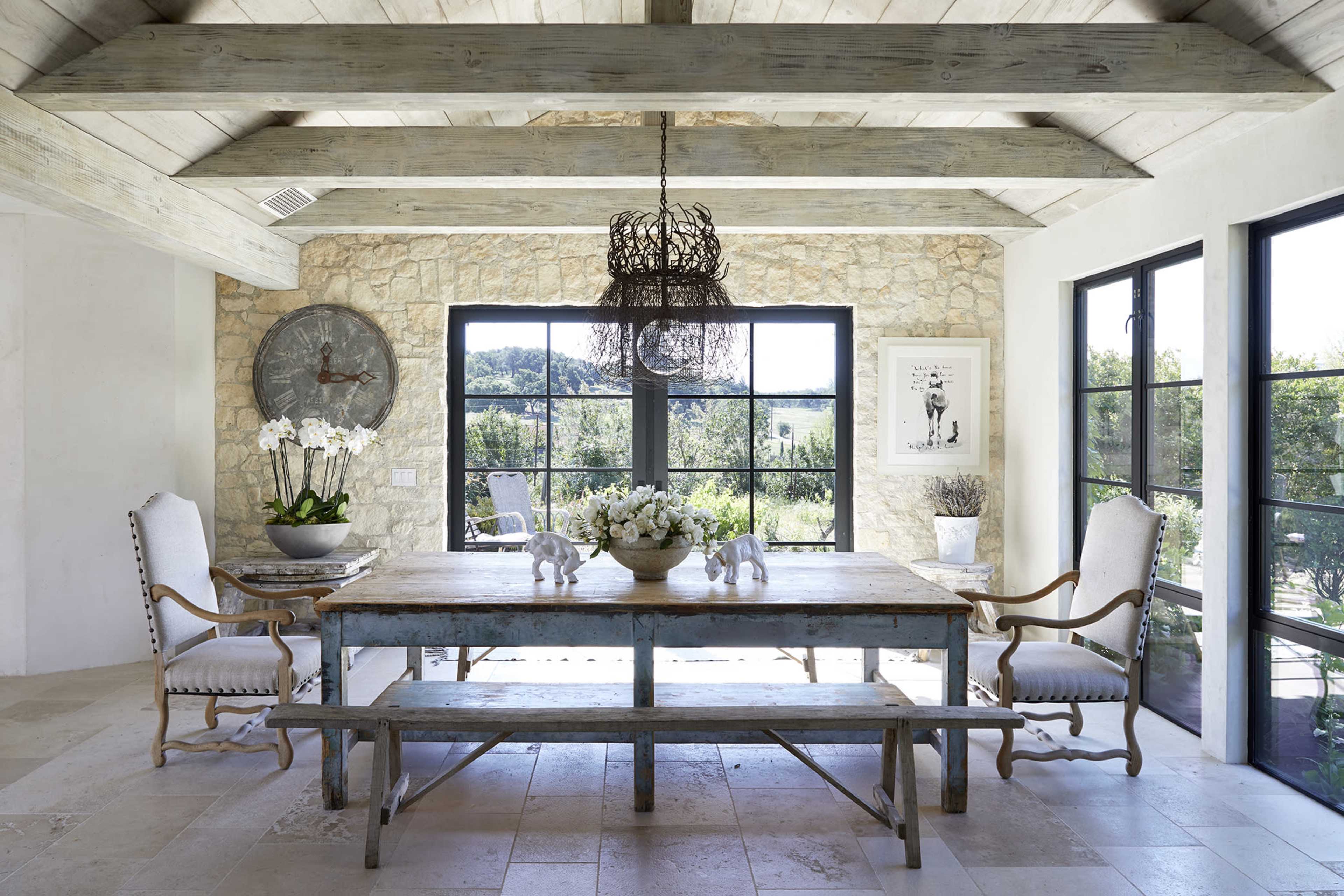 A rustic dining area featuring a wooden table, two chairs with armrests, a large clock on the wall, and large windows providing a view of the outdoors.