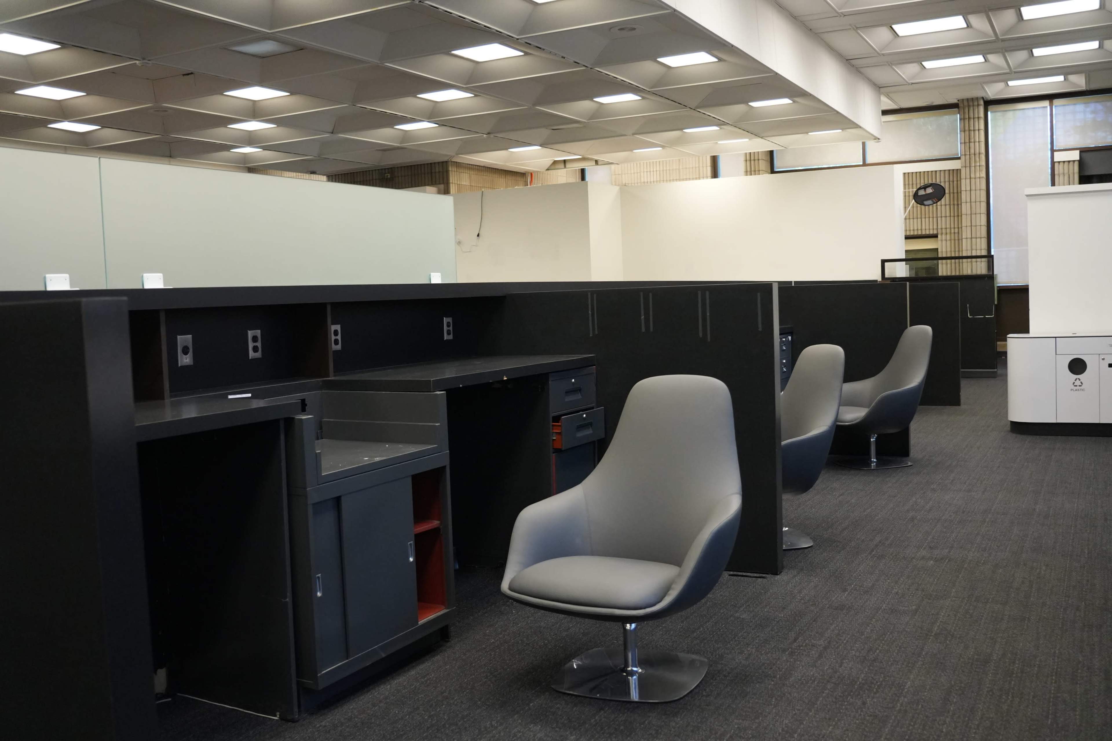 The image shows an empty office space with gray desks and chairs arranged in a row, featuring a carpeted floor and overhead lighting.