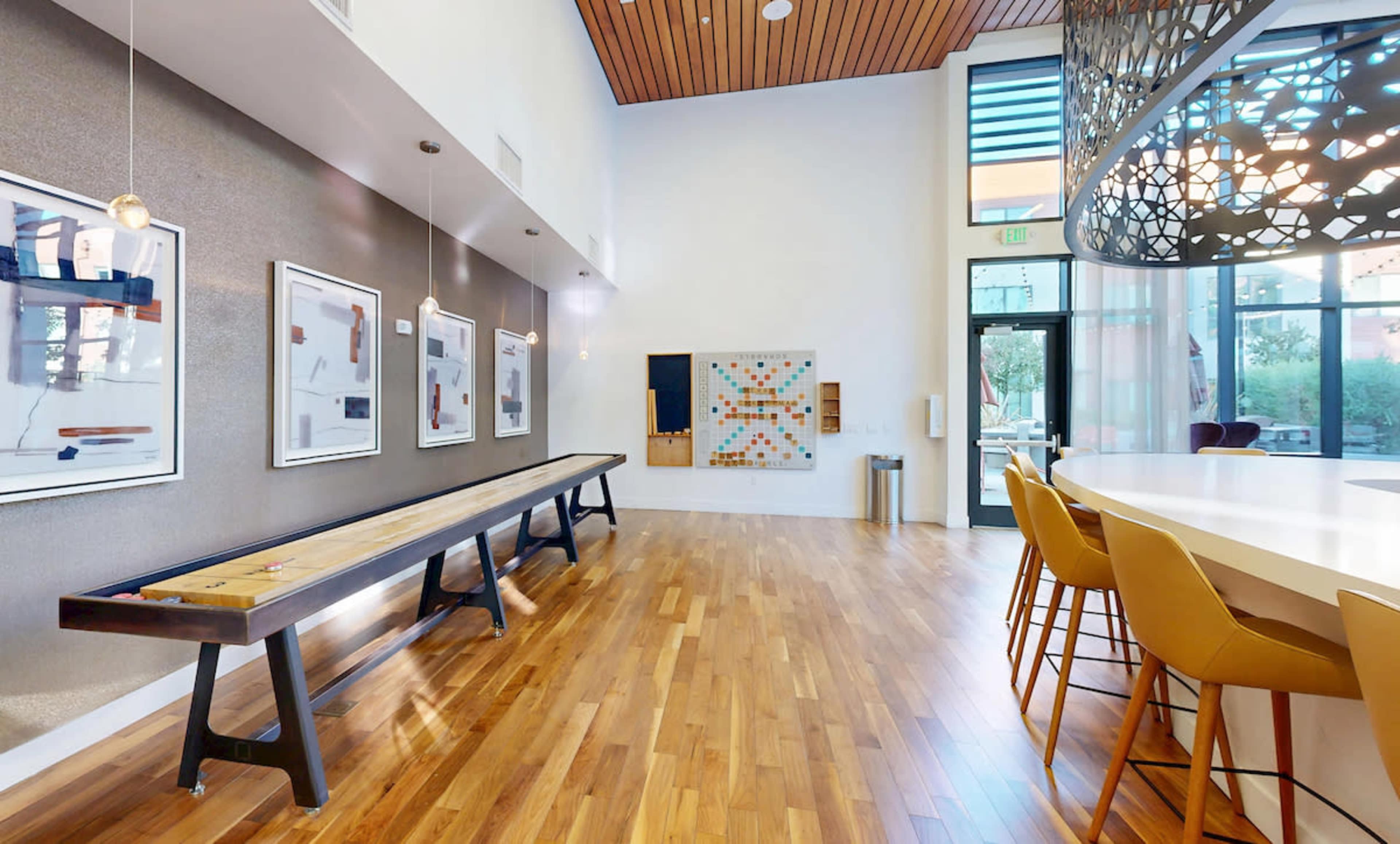 The image shows a spacious modern room with a wooden floor, featuring a long table, several framed artwork pieces on the wall, and a bar area with high chairs.
