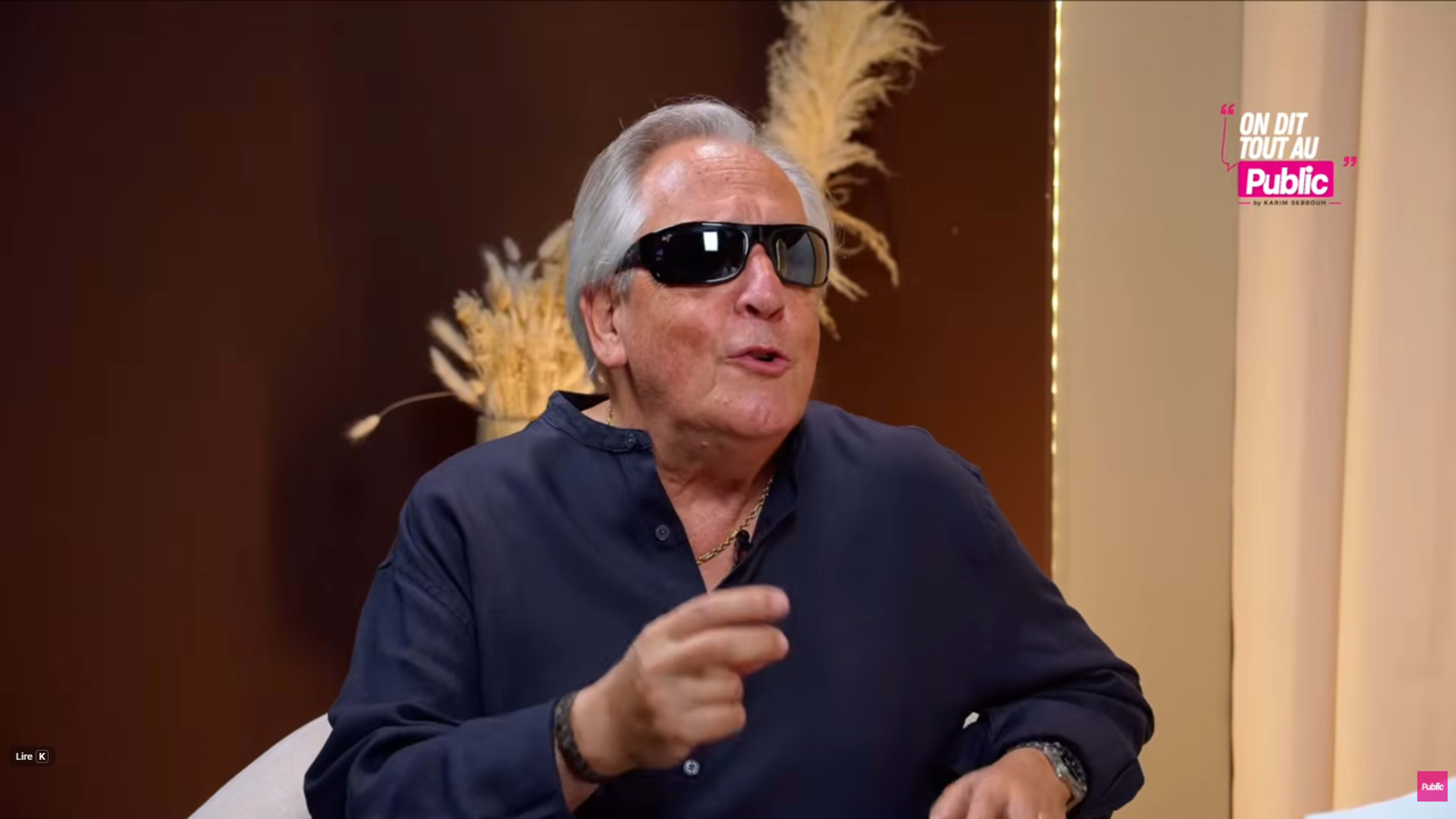 A man wearing sunglasses is speaking animatedly during an interview setting with decorative elements in the background.