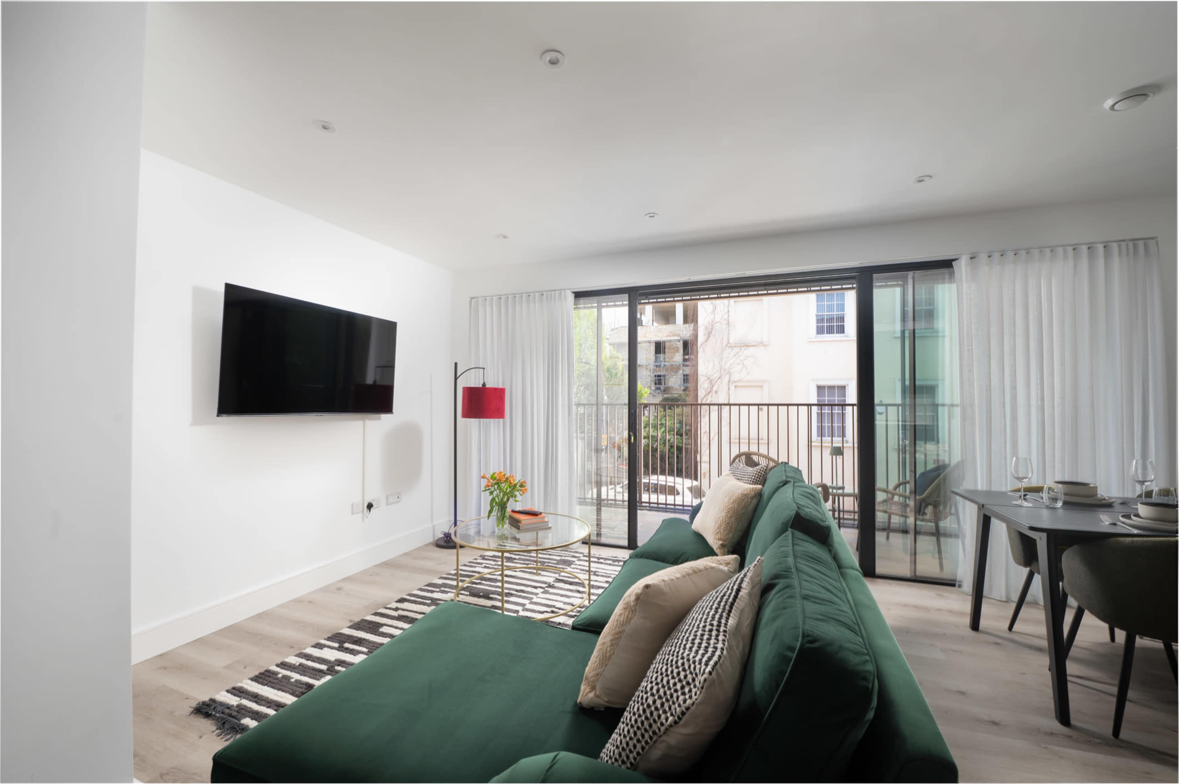A modern living room features a green sofa, a wall-mounted television, and a balcony with glass doors overlooking an outdoor space.