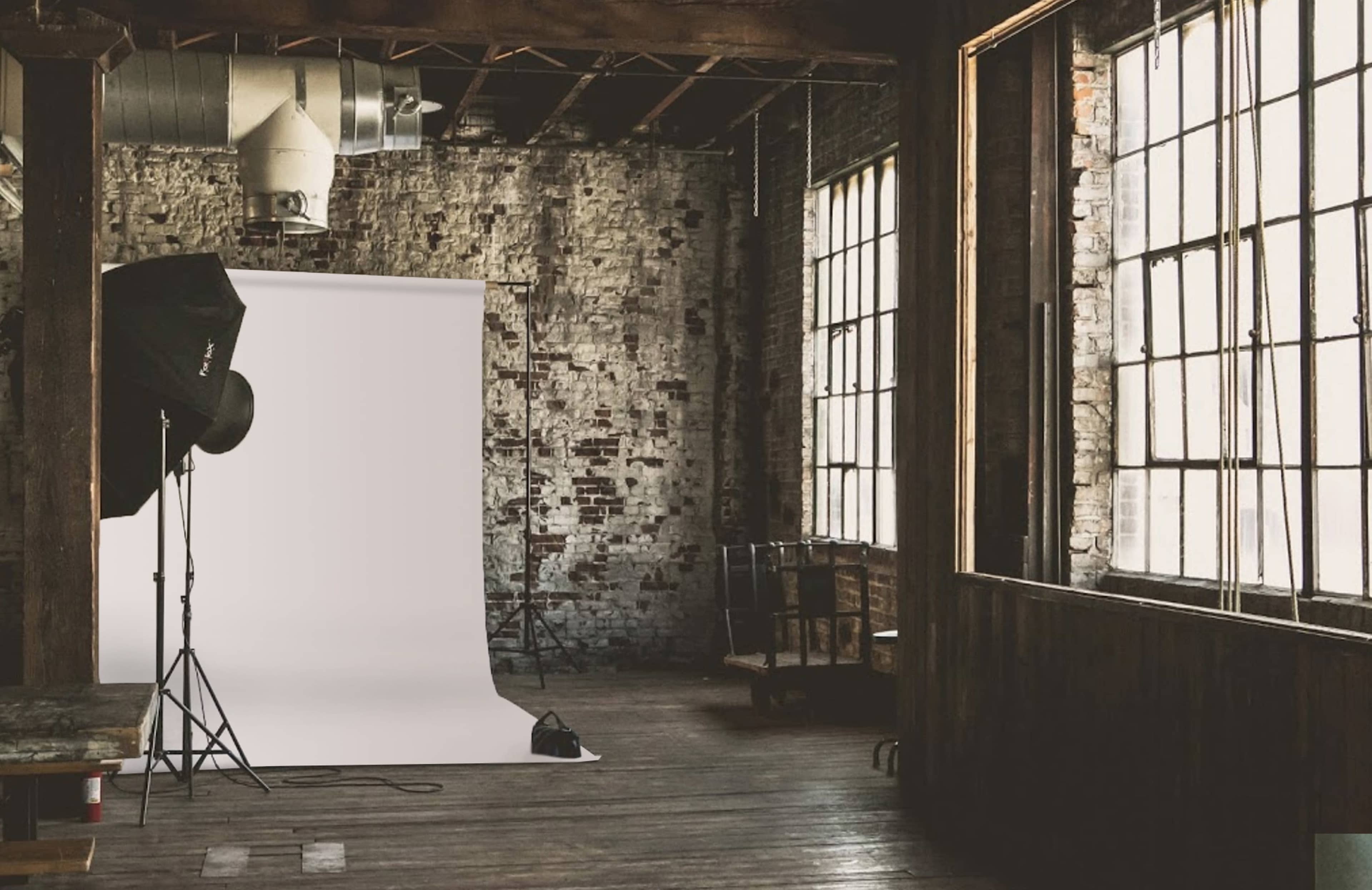 The image shows an empty industrial studio space with a white backdrop set up against a brick wall and large windows.