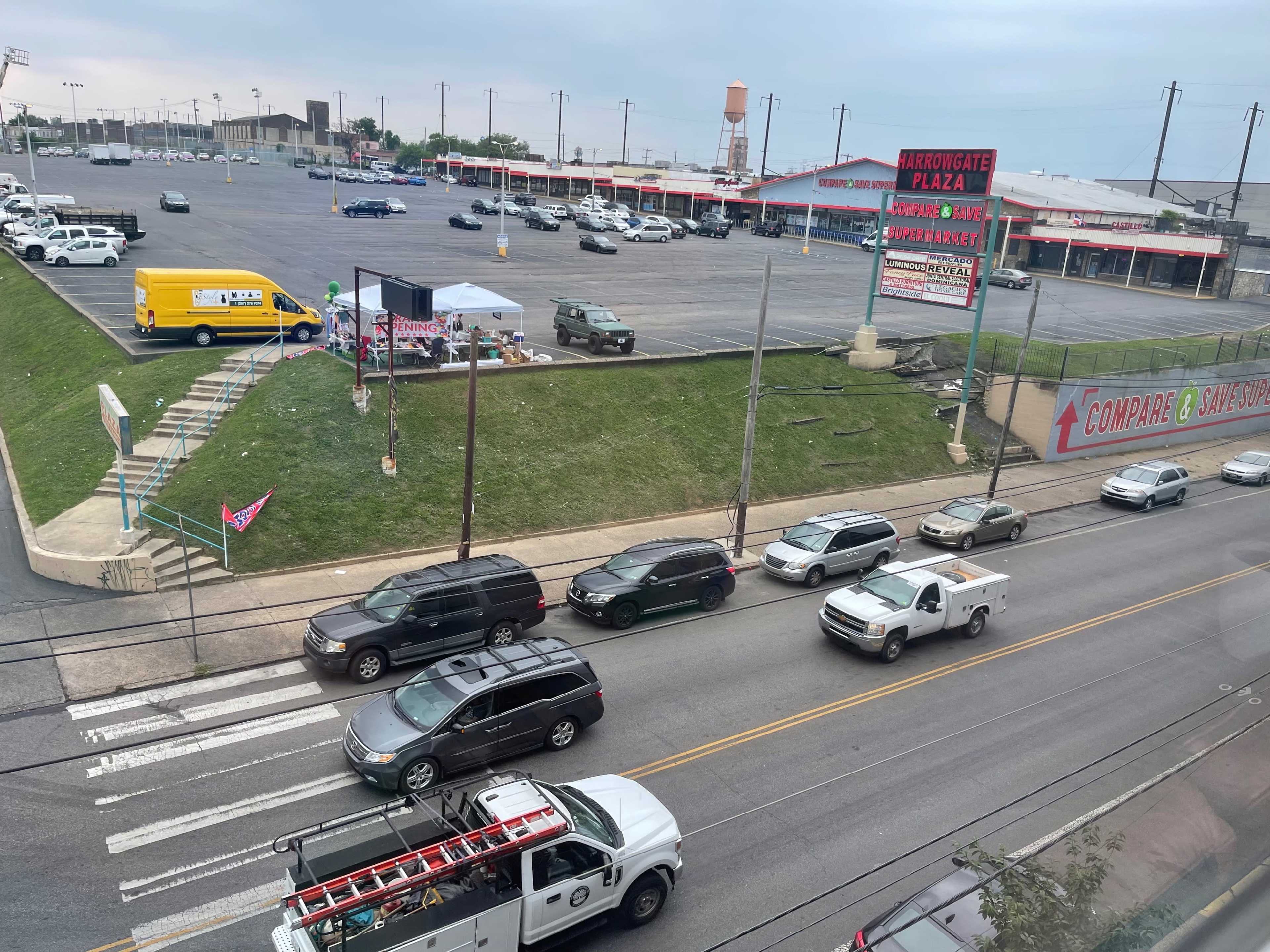 The image shows a busy street scene with multiple vehicles passing by a large parking lot and a nearby building displaying signage.
