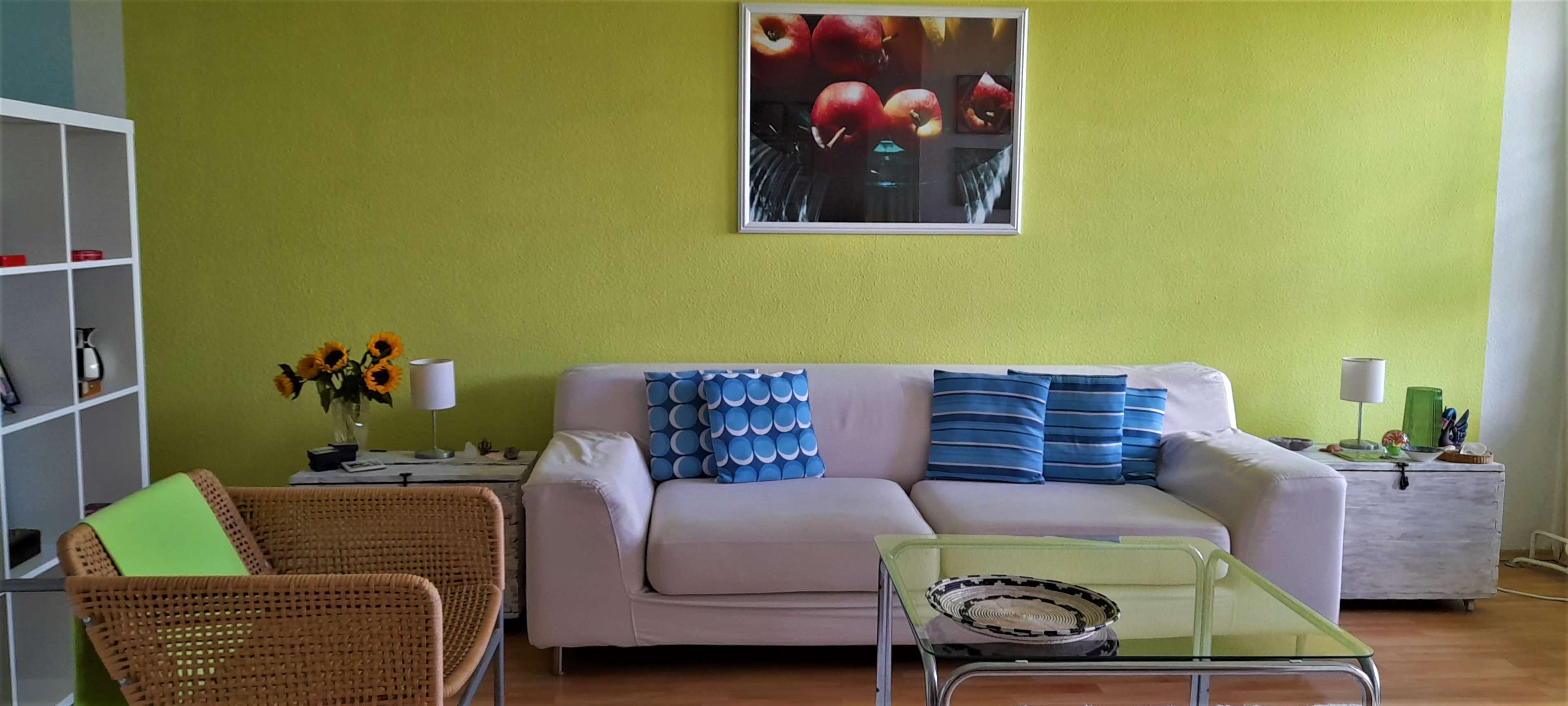 A living room features a white couch with blue and green cushions, a glass coffee table, and a vibrant green wall with a fruit-themed artwork.